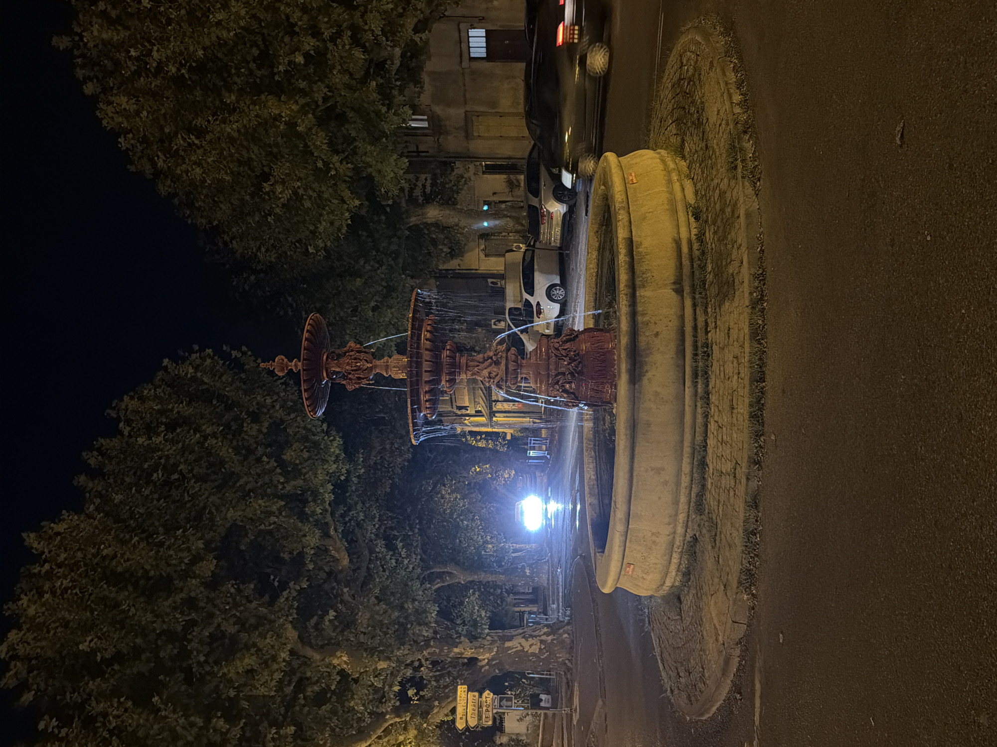 The image depicts a nighttime scene of a decorative fountain in the city of Viviers, France. The fountain is made of red, ornate metal and has water flowing from its upper tiers into a large stone basin. The basin is surrounded by a circular paved area, with a street curving around it. Several cars are visible on the street, with one car in the background having its headlights on, illuminating the wet road. Tall trees with dense foliage line the street behind the fountain, and buildings can be seen in the distance. A street signpost is visible to the left, indicating directions to "Theatre," "Le POSTE," and other locations. The overall atmosphere is serene and illuminated by streetlights and car headlights.