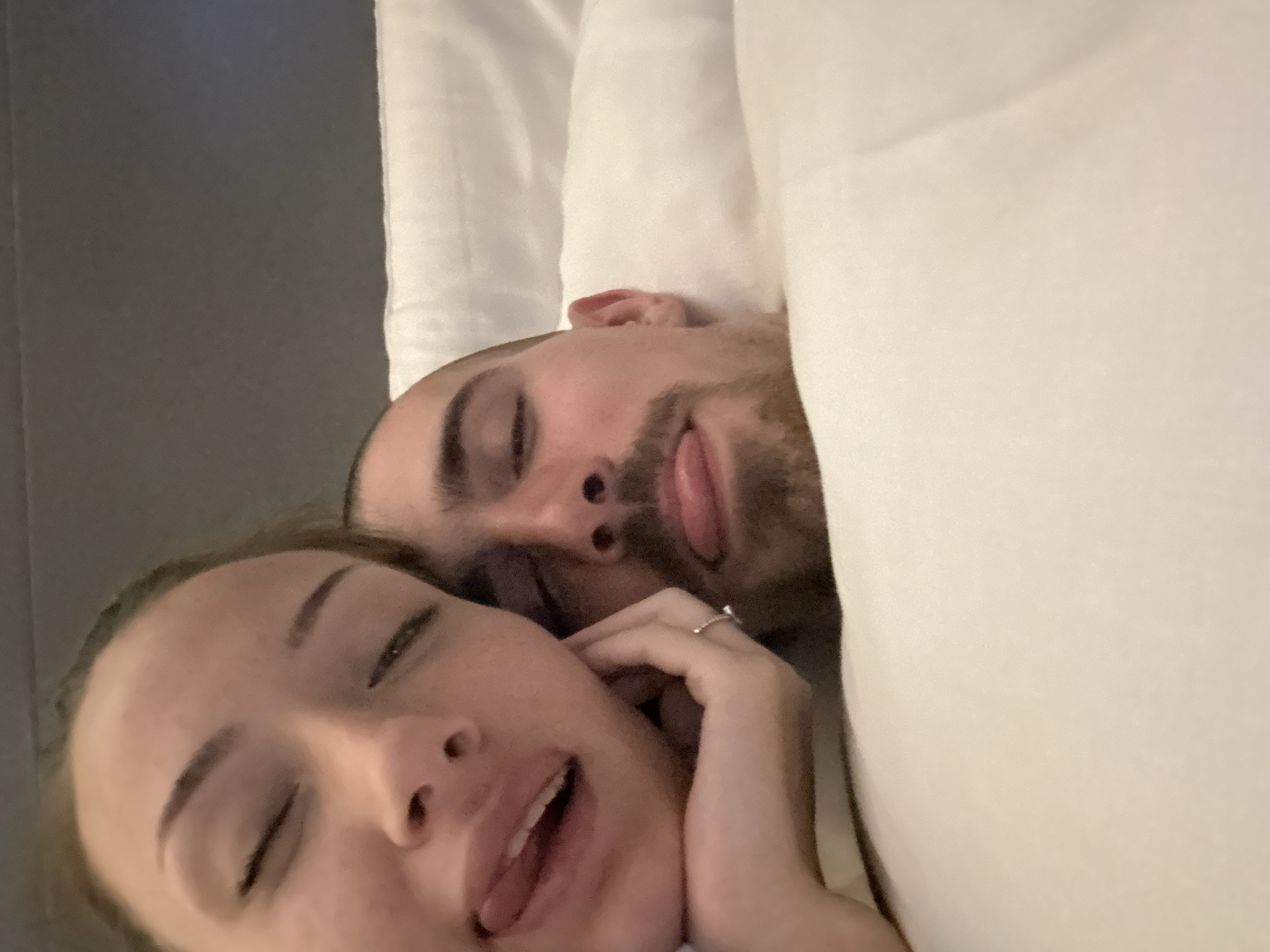 The image depicts two individuals, a female and a male, in a close-up, indoor setting. They are positioned horizontally, appearing to be lying on a bed with white pillows and bedding visible. The background consists of a dark, plain surface, possibly a headboard or wall.
The female is on the left, closer to the camera. She has light skin, dark hair partially visible, and her eyes are closed or heavily squinted. Her mouth is open, with her tongue slightly protruding. Her right hand, featuring a ring with a small stone on the ring finger, rests against her cheek/chin.
The male is positioned to the right of and slightly behind the female. He has light skin, short dark hair, and a stubble beard. His eyes are also closed or heavily squinted, and his mouth is open with his tongue slightly protruding, mirroring the female's expression.
Both individuals exhibit a playful or relaxed demeanor through their facial expressions. The interaction shows them in close proximity, sharing a similar pose.
The specific location within the city of Varna, Bulgaria, cannot be determined from the visual information provided in the image.