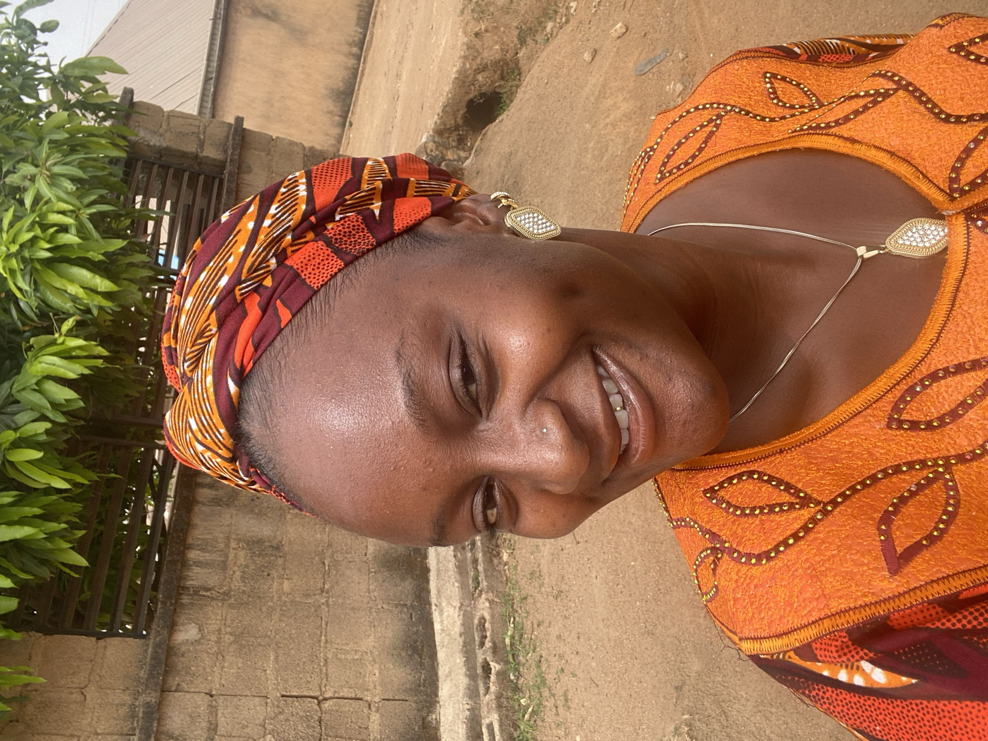 The image shows a smiling woman outdoors in Barakin Akawo, Nigeria. She wears a patterned headscarf and an orange dress with decorative beads. She has a silver necklace with a teardrop pendant and stud earrings. Her nose is pierced. The background includes greenery, a wall, and what appears to be a paved or unpaved path.