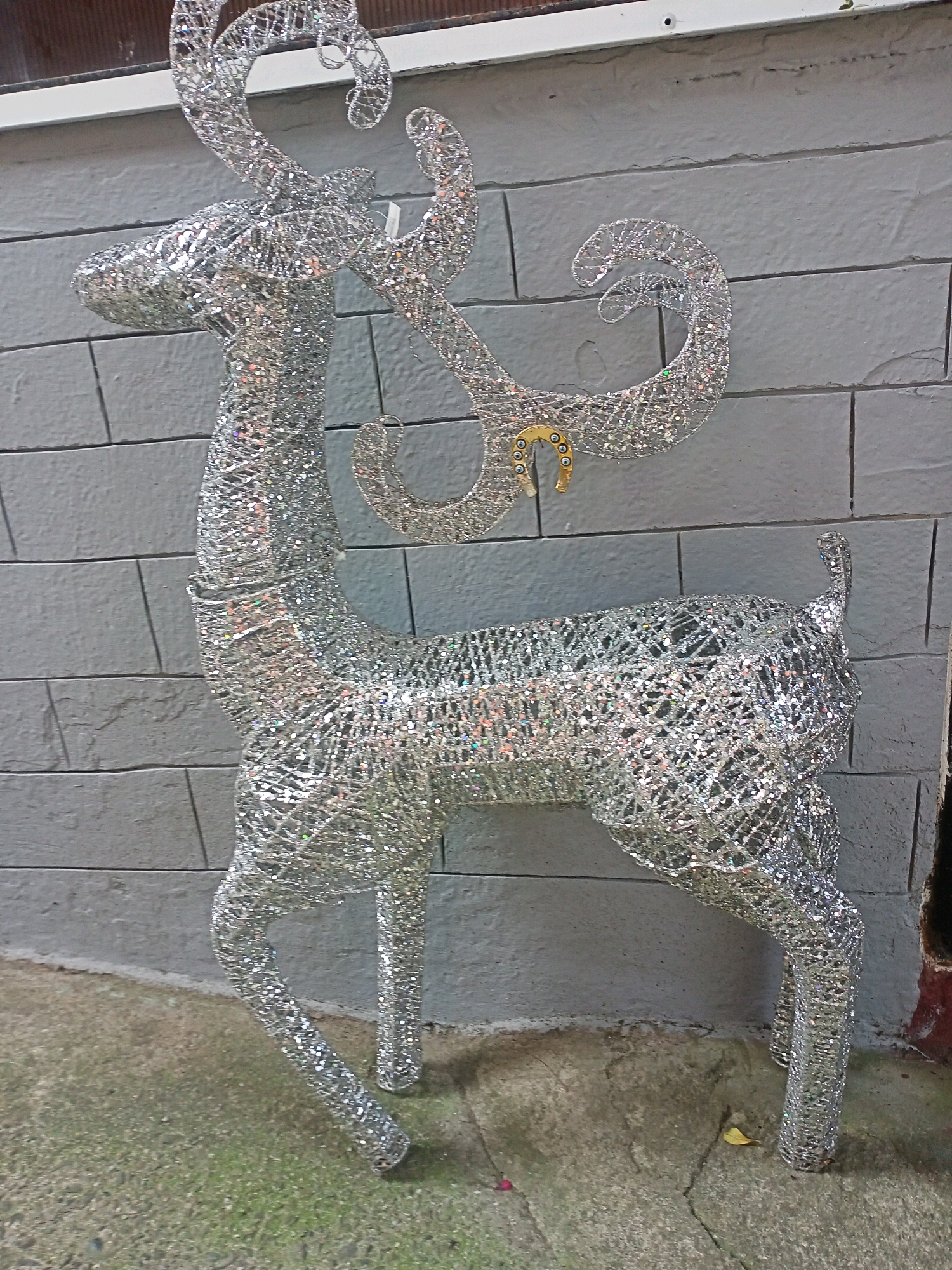 A silver, wire-frame reindeer decoration stands outside a grey, brick-patterned wall. The reindeer is facing left, with its head slightly turned towards the viewer. It is adorned with a small, gold horseshoe with blue accents hanging from its antlers. The ground in front of the reindeer is a mix of concrete and mossy patches. The scene appears to be located in Tsandrypsh, Abkhazia.