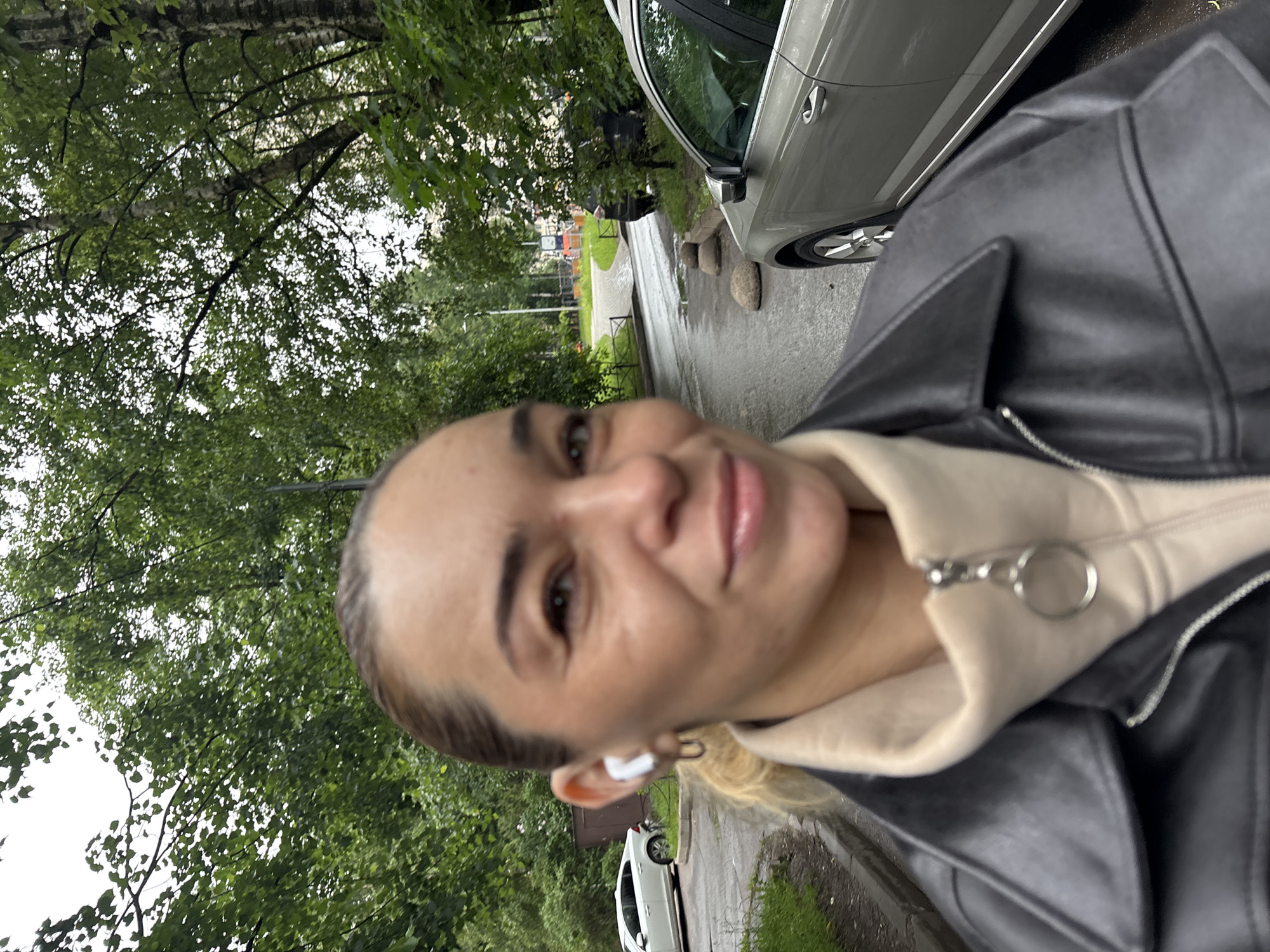 A woman is taking a selfie outdoors in Vsevolozhsk, Russia. She is wearing a black leather jacket over a tan turtleneck and white earbuds. She has blonde hair pulled back and is smiling at the camera. Behind her, a grey sedan is parked on a wet asphalt road. Trees and foliage line the street, and a white car is parked further down the road. The sky is overcast.