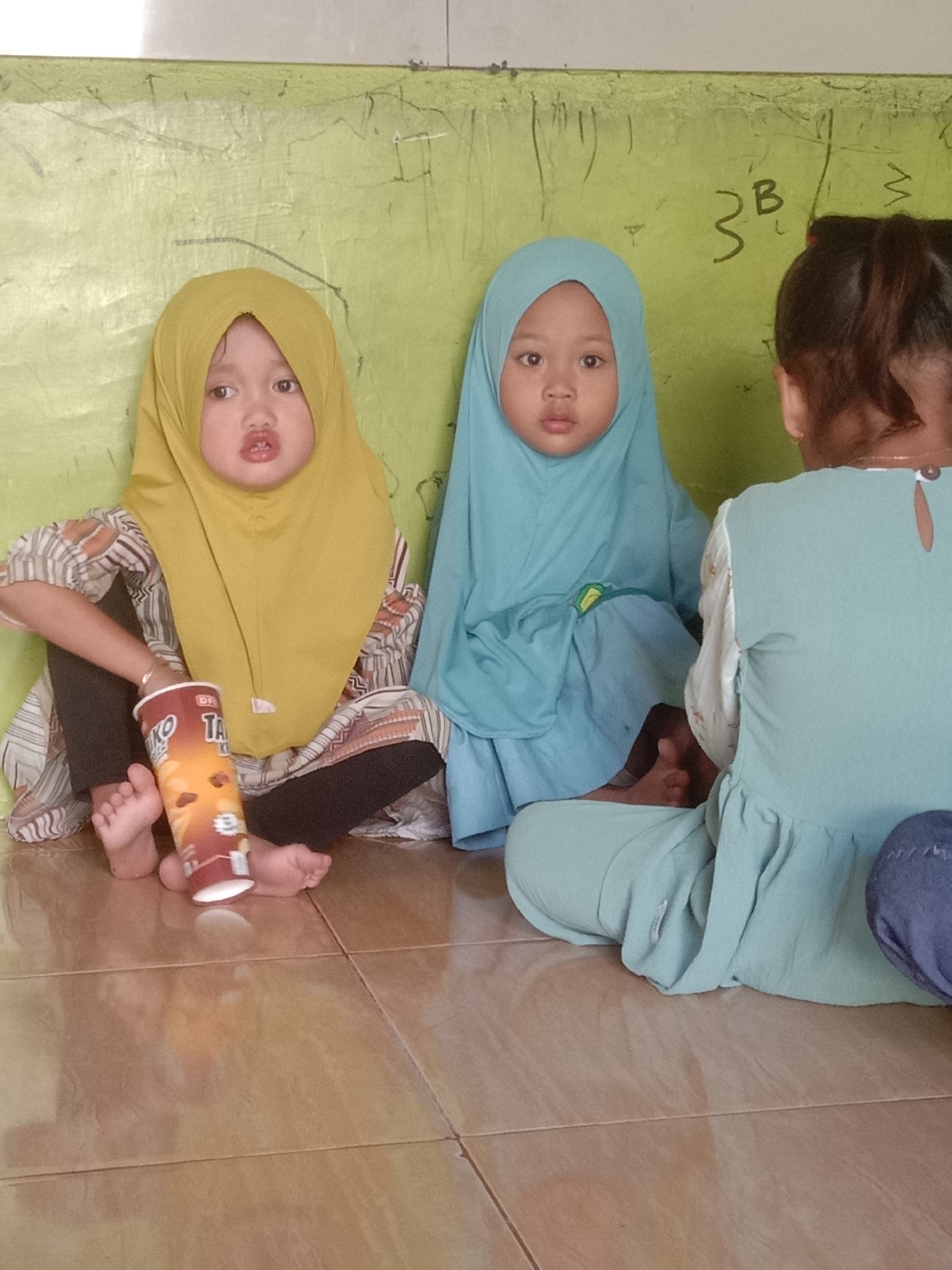 In a room with a green wall, two young girls wearing hijabs are sitting on the floor. The girl on the left wears a yellow hijab and a patterned shirt with black leggings. She holds a can of snacks. The girl to her right wears a blue hijab and a blue dress. A third person, seen from the back, wears a light green dress and sits to the right of the other two girls. The floor is tiled in a light brown color. The green wall has some markings on it, including what appears to be "3B" and "W". This scene appears to be in Tulungrejo, Indonesia.