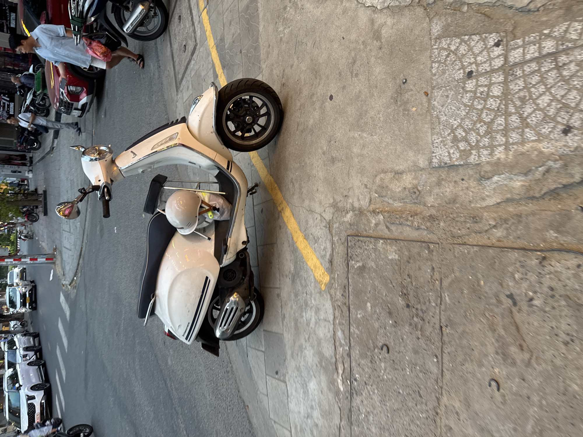 A white Vespa scooter with a black seat and a white helmet rests on a concrete sidewalk in Da Nang, Vietnam. A yellow line is painted on the sidewalk in front of the scooter. In the background, a red SUV is parked and a man in a light blue short-sleeved shirt and white shorts walks past, holding a pink plastic bag. Other vehicles, including white SUVs and motorcycles, are visible on the street. The scene appears to be a street in a city, with buildings and shops lining the road.