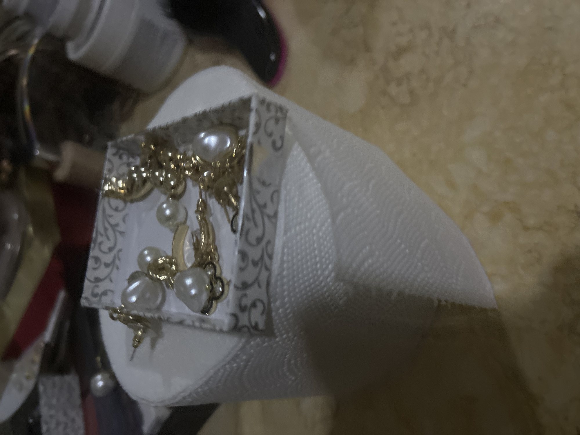 A close-up shot shows a small, square, decorative box filled with gold-colored jewelry featuring pearl-like beads. The box rests on a roll of white toilet paper. Other miscellaneous objects are vaguely visible in the blurred background. The scene's location cannot be determined from this image.
