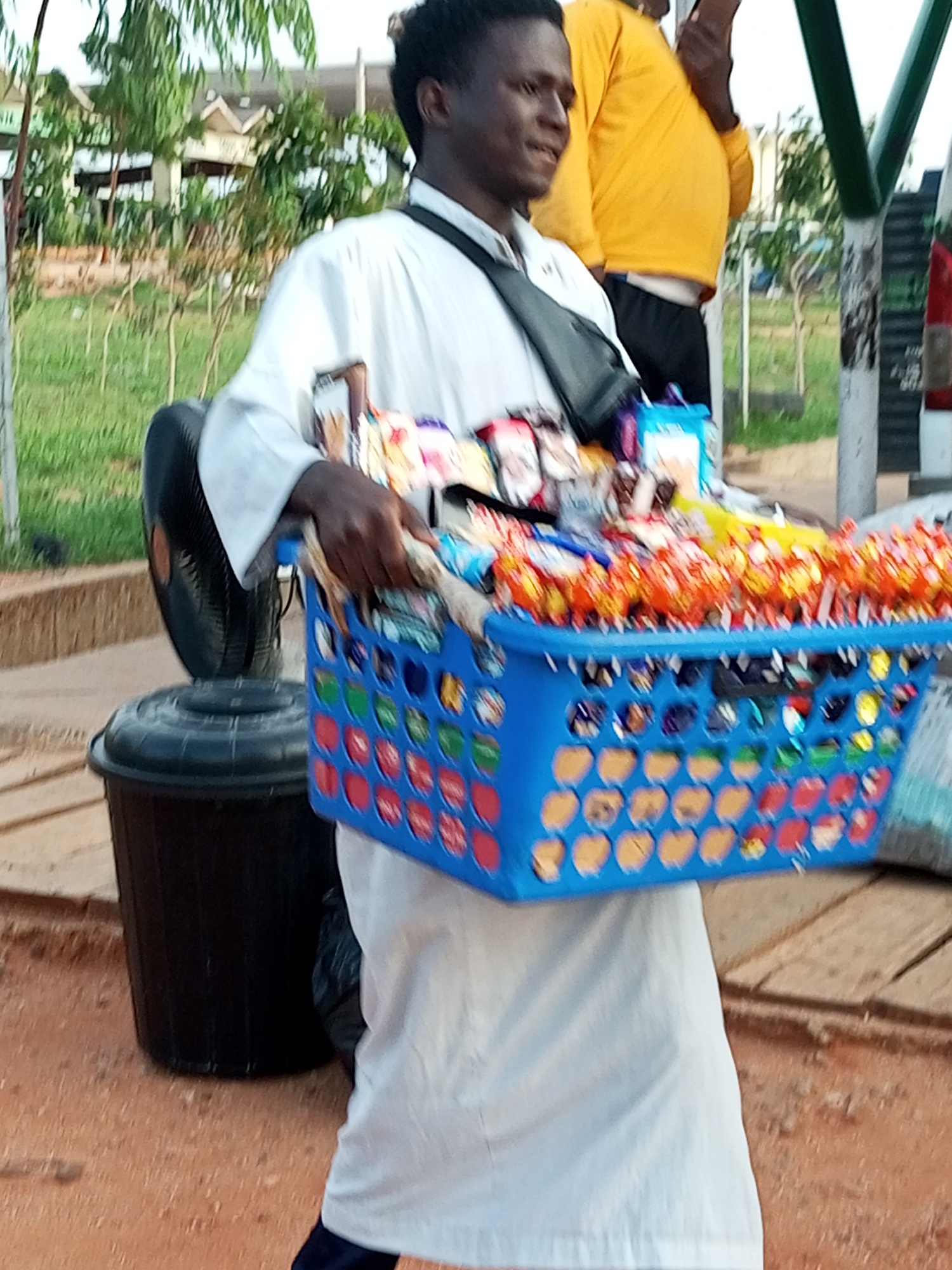 In Gombe, Nigeria, a young Black male, wearing a white robe and a dark crossbody bag, stands on a reddish-brown dirt surface. He is holding a blue plastic basket filled with various bagged snacks and candies, some of which are individually wrapped in orange and yellow packaging. To his left, a black electric fan is positioned on top of a large black cylindrical bin. In the background, there are trees, some buildings with beige walls and angled roofs, and a green metal pole with a white and black striped section. Another individual, wearing a bright yellow long-sleeved shirt and black pants, is visible behind the primary subject, holding a dark object, possibly a phone, to their ear. The overall scene suggests a street vending environment.