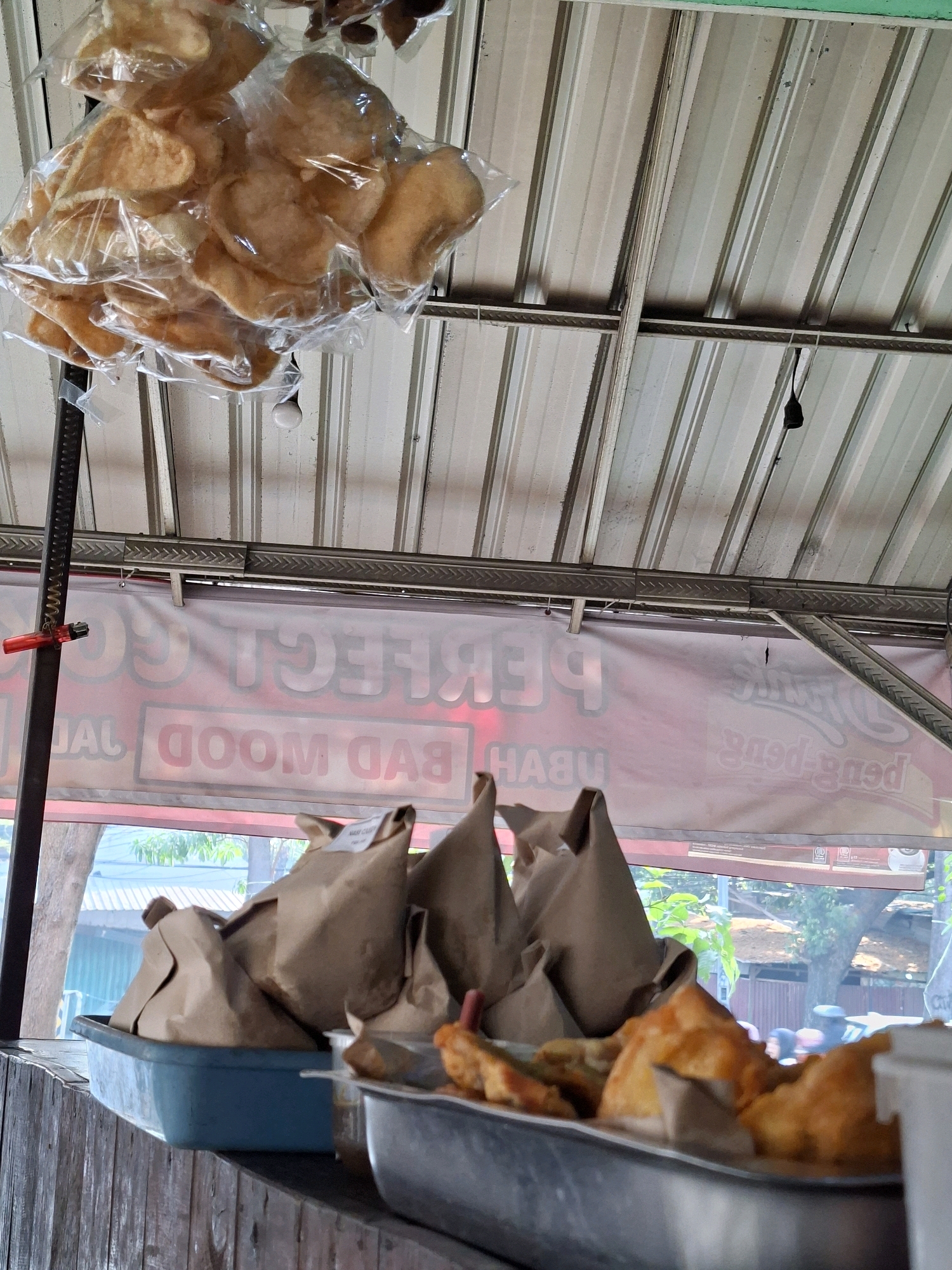 In Surabaya, Indonesia, a street food stall operates under a corrugated metal roof. A bag of golden-brown crackers, possibly krupuk, is suspended in plastic packaging from the ceiling. Below, a pale pink banner with blurry red lettering, potentially indicating "PERFECT GOOD MOOD," stretches across the stall. A blue plastic tub contains several brown paper-wrapped parcels, presumably containing food items. In the foreground, a metal tray holds more fried food items, including what appear to be spring rolls and other savory snacks. The stall is situated outdoors, with glimpses of trees and buildings visible through a window or opening. The overall scene depicts a bustling, everyday food vendor in a typical Indonesian urban setting.