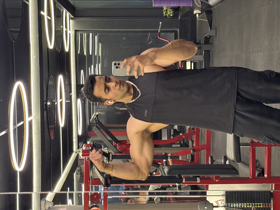 An adult male takes a mirror selfie in a modern gym located in Kolkata, India. The male subject, appearing to be of South Asian descent, is positioned centrally, wearing a black H&M sleeveless tank top, black cargo shorts, a gold-colored neck chain, and a gold-colored bracelet on his left wrist. He has short, dark hair, light facial stubble, and white wireless earbuds in both ears. He holds a light-colored smartphone with multiple cameras in his right hand, pointed towards the mirror, capturing his reflection. His left arm is bent, and he is flexing his bicep.
The gym environment features black walls and ceiling with several circular, gold-rimmed LED lights and linear strip lighting. Red and black gym equipment, including a cable machine with a weight stack, is visible in the background, primarily on the left. A black padded bench and dumbbells are also present. A purple foam roller and a green potted plant are visible on the right side of the background. A partial reflection of another individual is faintly visible in the far-left background. The floor is covered with black gym mats.