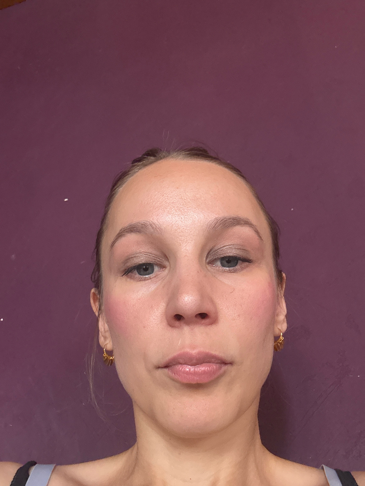 A woman is pictured from the chest up, against a plain purple background. Her head is tilted slightly back, and she is looking directly at the camera. She has fair skin with some visible pores and light blush applied to her cheeks. Her eyebrows are naturally shaped and light brown, and her eyelids are adorned with a subtle shimmer eyeshadow. Her eyes are open, revealing blue-gray irises, and her lashes are dark. Her nose is straight, and her lips are full and painted with a neutral, glossy lipstick. She is wearing small, gold hoop earrings with dangly charms. The background is a solid, deep purple color, with no discernible features or patterns. The lighting appears to be soft and diffused, casting no harsh shadows. The woman's hair is pulled back, with strands visible around her face. She is wearing a grey and black tank top. There are no specific landmarks or details in the image that would allow for the identification of a location in Überherrn, Germany.
