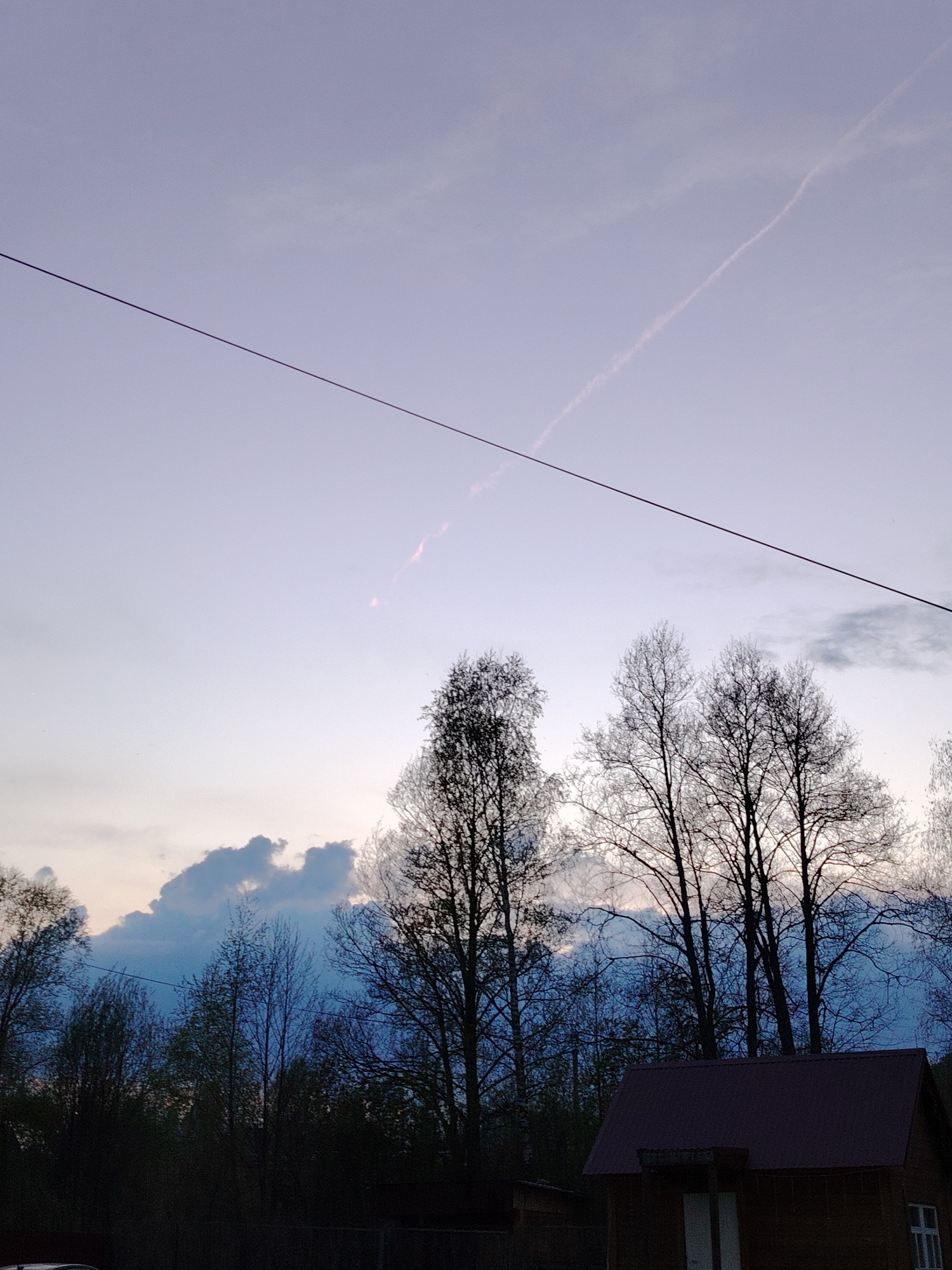 The image depicts a view of the sky at dusk in Malyi Bukor, Russia. The sky is a light purplish-blue with a faint contrail bisecting the upper right quadrant. A dark, horizontal power line crosses the upper portion of the frame. In the middle ground, silhouettes of bare trees stand against a backdrop of blue-tinged clouds. In the foreground, the roof of a small wooden building is visible. Some vegetation is also seen at the bottom of the image, suggesting a wooded or residential area.