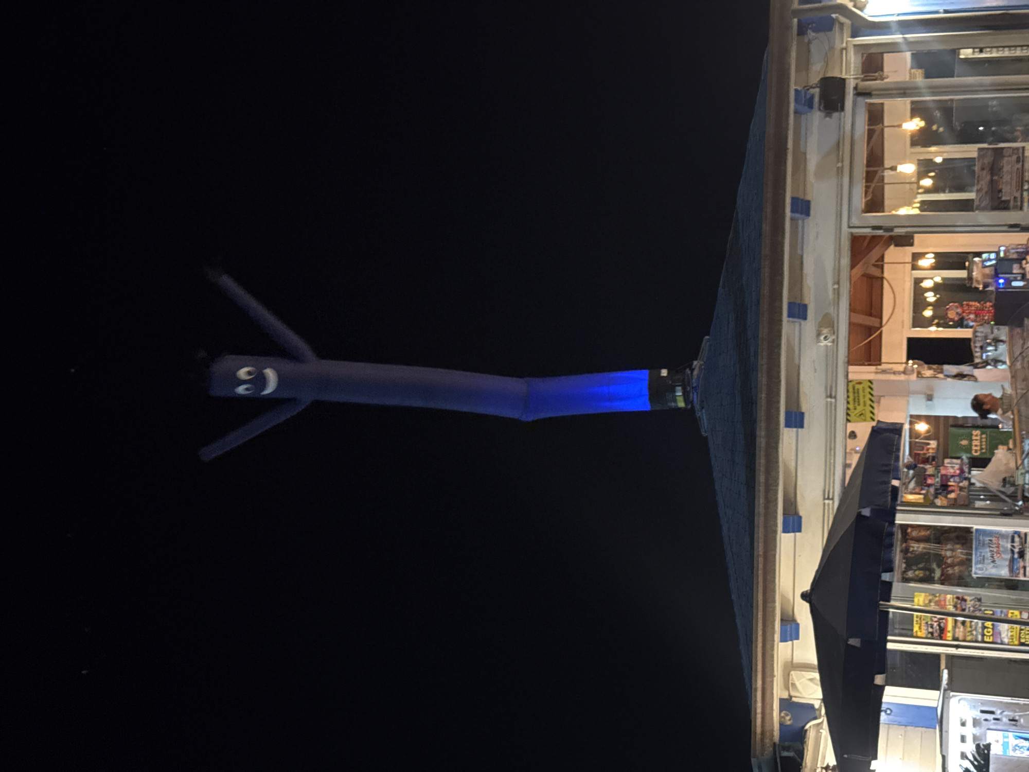 An inflatable tube dancer, colored deep blue with a smiling face, is positioned vertically above a low-rise building with a dark blue roof, illuminated by blue light at its base. The building appears to be a shop or kiosk, with a large black umbrella in front of its glass storefront. Various items are visible inside the shop, including shelves stocked with products and promotional posters. A woman is partially visible inside the shop on the right, and an espresso machine is visible on the left. The scene is lit with artificial lights, suggesting it is nighttime. The building is located in Isola delle Femmine, Italy.