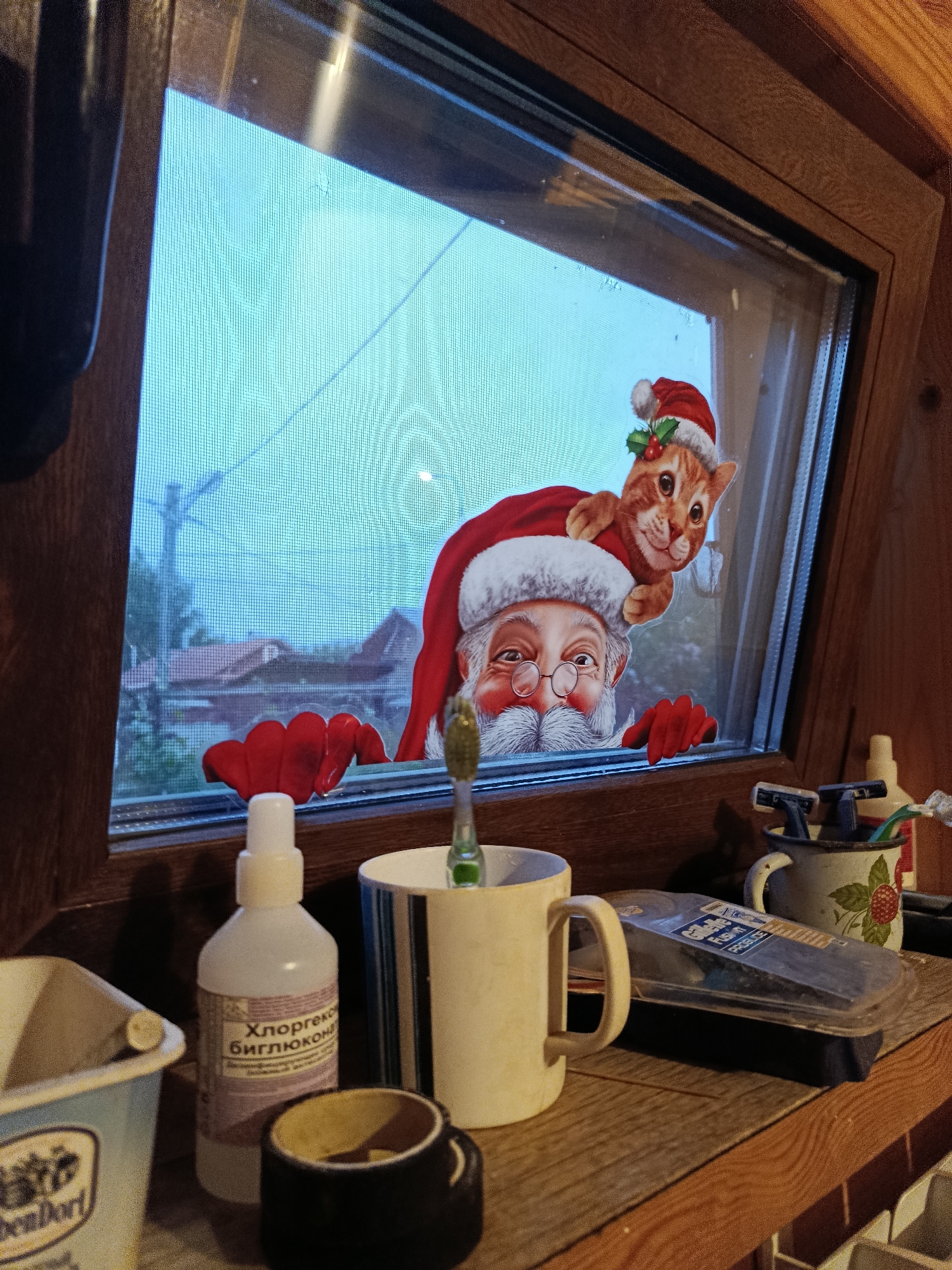 A close-up, interior shot displays a wooden shelf with various items. On the left, a light blue plastic container with a white label partially visible. Next to it is a white plastic bottle with a label in Russian that reads "Хлоргексидин Биглюконат" (Chlorhexidine Bigluconate). To the right of that, a dark rimmed object, possibly a small bowl or ashtray, sits on the shelf. Further right, a beige ceramic mug with a handle holds a green toothbrush. Behind the mug, a metallic cup with a red berry design contains two disposable razors. A clear plastic container with a blue and silver label is also present on the shelf. In the background, a window with a screen reveals an outdoor scene of houses and trees under a hazy sky. A sticker depicting Santa Claus and a cat in a Santa hat is affixed to the window. The overall impression is of a bathroom or utilitarian area within a home.