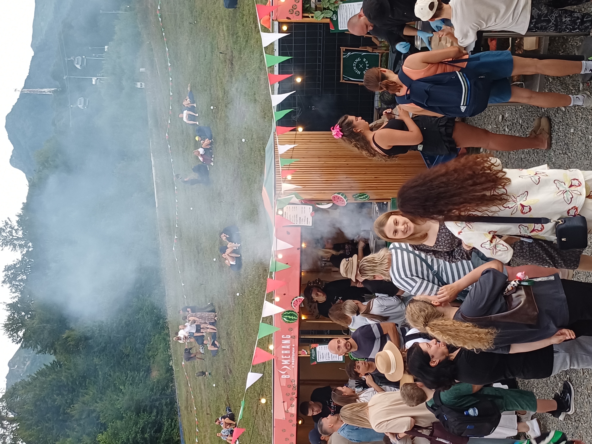 The image captures a bustling outdoor scene at a festival or market in Estosadok, Russia, likely during the warmer months given the attire of the attendees. In the foreground, a group of people are gathered around a food stall adorned with colorful pennants and fruit-themed decorations, including watermelons. The stall appears to be serving food, with staff members actively engaged in preparation. A noticeable amount of smoke or steam is rising from the stall, adding to the atmosphere.
Behind the food stall, the terrain slopes upwards, covered in grass and dotted with people relaxing or socializing. In the background, majestic mountains rise, partially obscured by trees and a significant amount of smoke or mist. Cable cars are visible ascending the mountain, suggesting the area is a popular tourist destination or resort, likely associated with a ski area that operates during non-winter seasons.
The attendees are dressed in casual summer clothing, indicating a relaxed and informal event. The overall impression is one of a lively outdoor gathering with food, entertainment, and scenic mountain views.