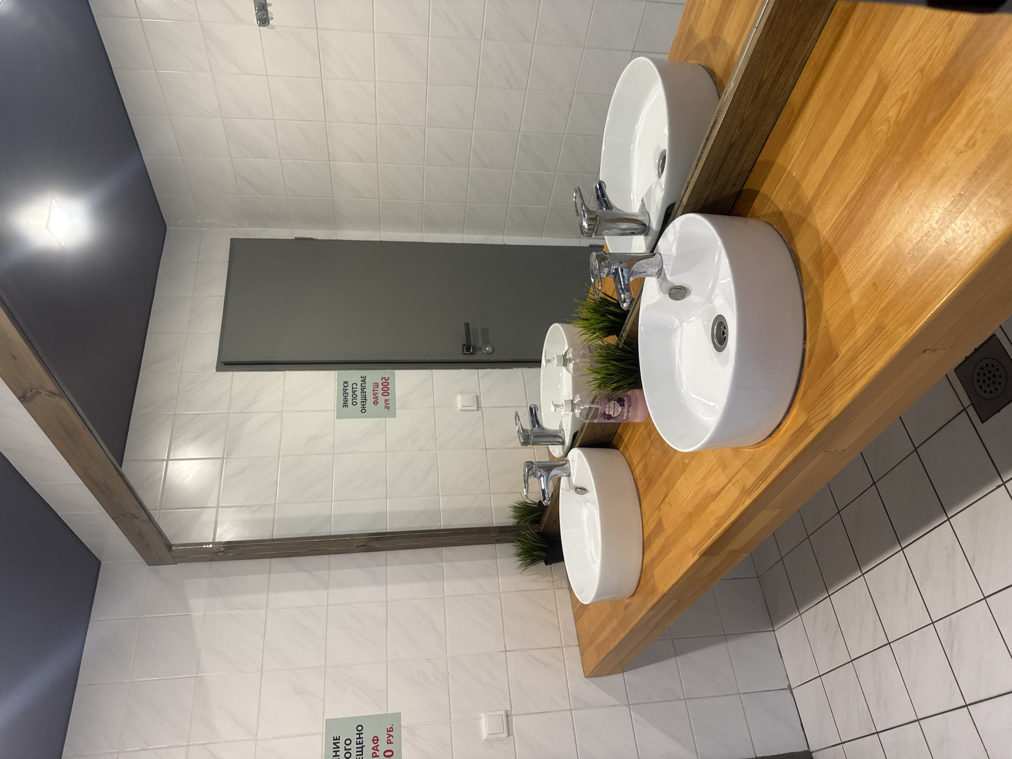 The image displays a clean, modern public washroom or restroom interior, likely located in Tobolsk, Russia. The primary subjects are a series of three white, circular, vessel-style sinks set upon a long, light-colored wooden countertop. Each sink is fitted with a chrome faucet.
A large mirror with a wooden frame occupies the wall behind the sinks, reflecting the opposite side of the room. In the reflection, a gray door with a handle is visible on the left, along with a section of a dark gray wall featuring a bright light fixture. The walls are predominantly covered in white square tiles with light gray grout. The floor is also tiled with white squares, and a dark circular drain is visible in the lower right corner of the frame.
On the wooden counter, between two of the sinks, there is a small artificial green plant and a clear pump soap dispenser. No specific activity is taking place in the image; it is an unoccupied interior shot. The lighting is artificial, indicating an indoor setting.
Notable details include two warning signs with text in Russian. Reflected in the mirror, an inverted sign reads: "ВНИМАНИЕ! ШТРАФ 2000 РУБ. ЗА ПОРЧУ ИМУЩЕСТВА СТОЛО" (likely "СТОЛОВОЙ," meaning "Attention! Fine 2000 rubles for damage to cafeteria property"). A similar, smaller sign is directly visible on the tiled wall below the mirror, reading: "ВНИМАНИЕ! ШТРАФ 500 РУБ. ЗА ПОРЧУ ИМУЩЕСТВА" (Attention! Fine 500 rubles for damage to property). These signs suggest the washroom is part of a public or commercial establishment, possibly a cafeteria.