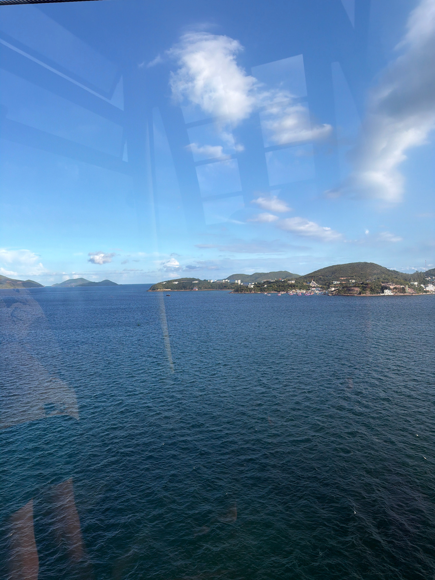 The image shows a wide expanse of deep blue ocean under a bright blue sky with scattered white clouds. The ocean surface has a gentle ripple. In the distance, a lush green coastline with hills is visible, dotted with buildings and what appear to be boats. The shoreline is lined with trees and some structures. A cable is visible at the top of the image, and reflections of a window frame are present in the foreground, suggesting the photo was taken from inside a vehicle or building. The scene depicts a coastal view, likely in Nha Trang, Vietnam, given the characteristic tropical landscape and the presence of numerous colorful boats, which are typical of Vietnamese fishing villages and tourist areas.