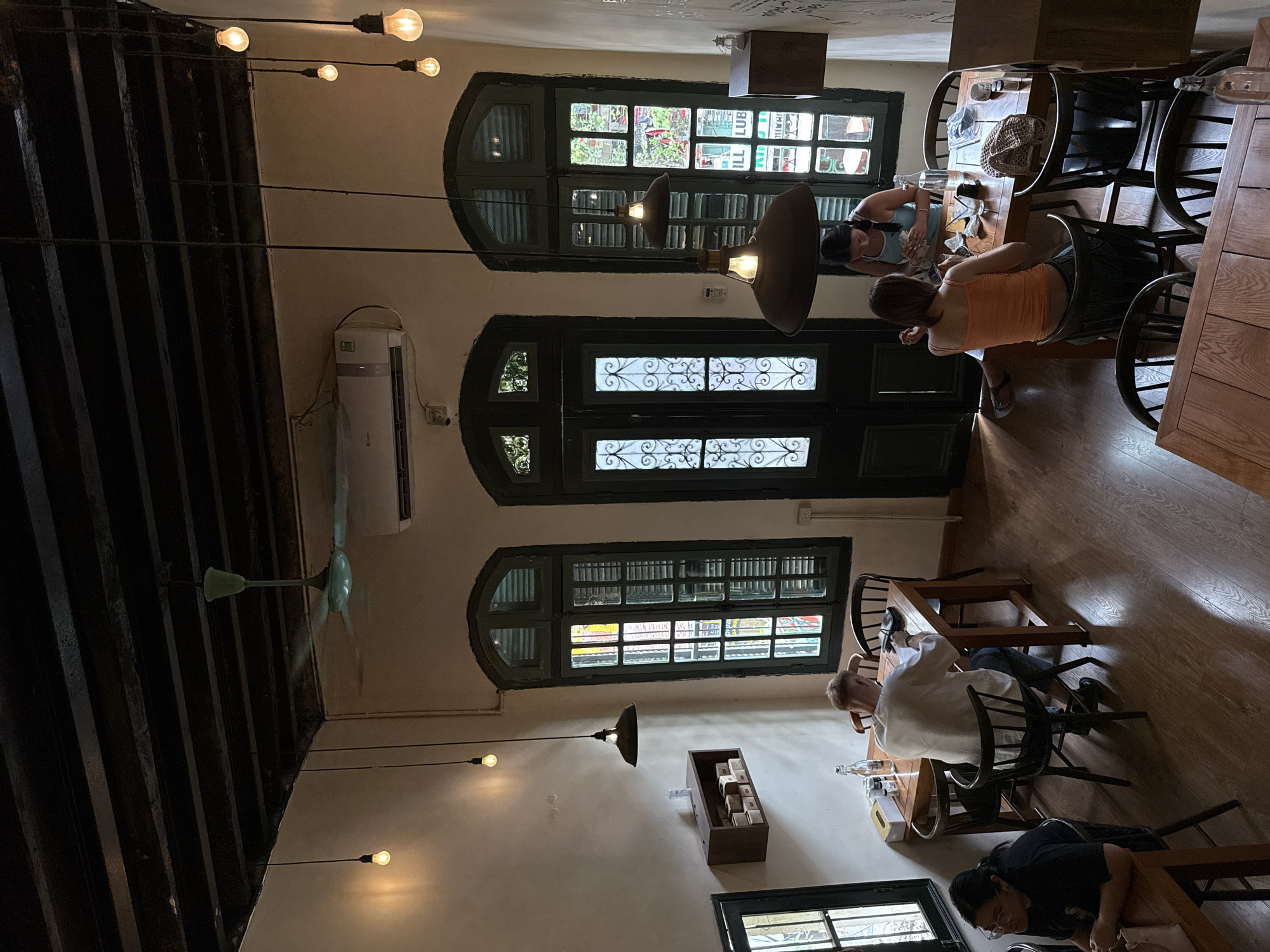 This image depicts the interior of a dimly lit cafe in Hanoi, Vietnam, characterized by exposed wooden beams on the ceiling and large, dark-framed windows with intricate designs. A ceiling fan is visible, as is an air conditioning unit. Several pendant lights hang from the ceiling, illuminating the space.
In the foreground, a woman with glasses wearing a black t-shirt sits at a wooden table, facing left. To her right, a man with blonde hair, wearing a white t-shirt, sits at a rectangular wooden table and appears to be writing or looking at something on the table. Behind him, a woman in an orange tank top and jeans sits at the same table, facing away from the camera, and a woman in a blue tank top is seated opposite her. They appear to be engaged in conversation or eating.
The cafe has wooden floors and tables. The walls are a light beige color, and there are dark green window frames and doors. A wooden shelf with boxes is mounted on the left wall, and a dark wooden cabinet is situated on the right wall. Bottles of water, glasses, and other items are on the tables. The overall ambiance suggests a relaxed, possibly vintage, setting for dining or socializing.