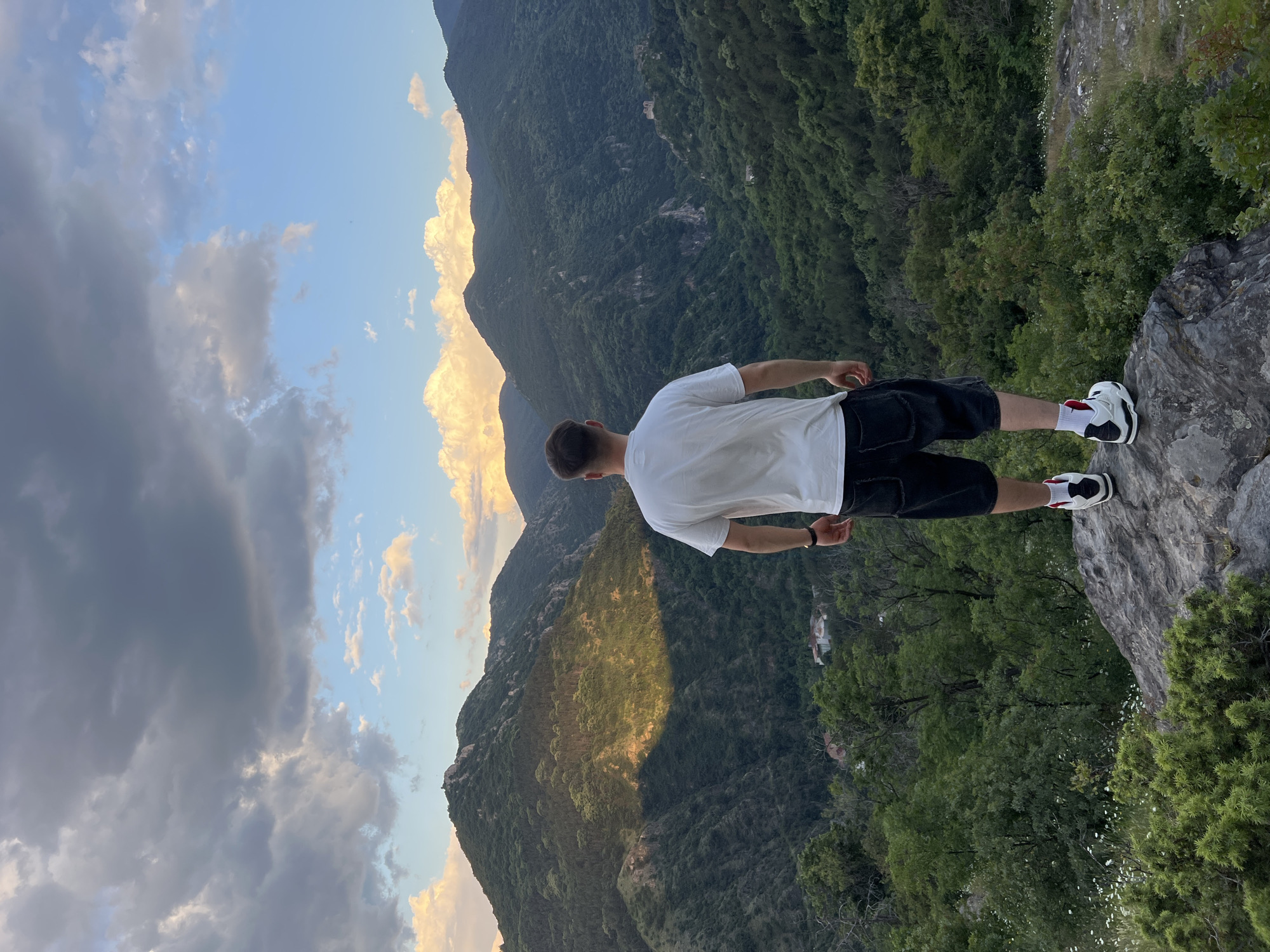 A person stands on a rocky outcrop overlooking a mountainous landscape. The individual is seen from the back, wearing a white t-shirt, dark shorts, and black and white sneakers with red socks. The landscape includes mountains covered in trees and foliage. The sky is visible with light blue interspersed with light and dark clouds, with golden light reflecting off some mountain peaks. The picture appears to be taken at an angle, slightly tilted, with the person and the immediate foreground situated towards the right side of the frame. The specific location in Asenovgrad, Bulgaria, is not evident from the visual details provided in the image.
