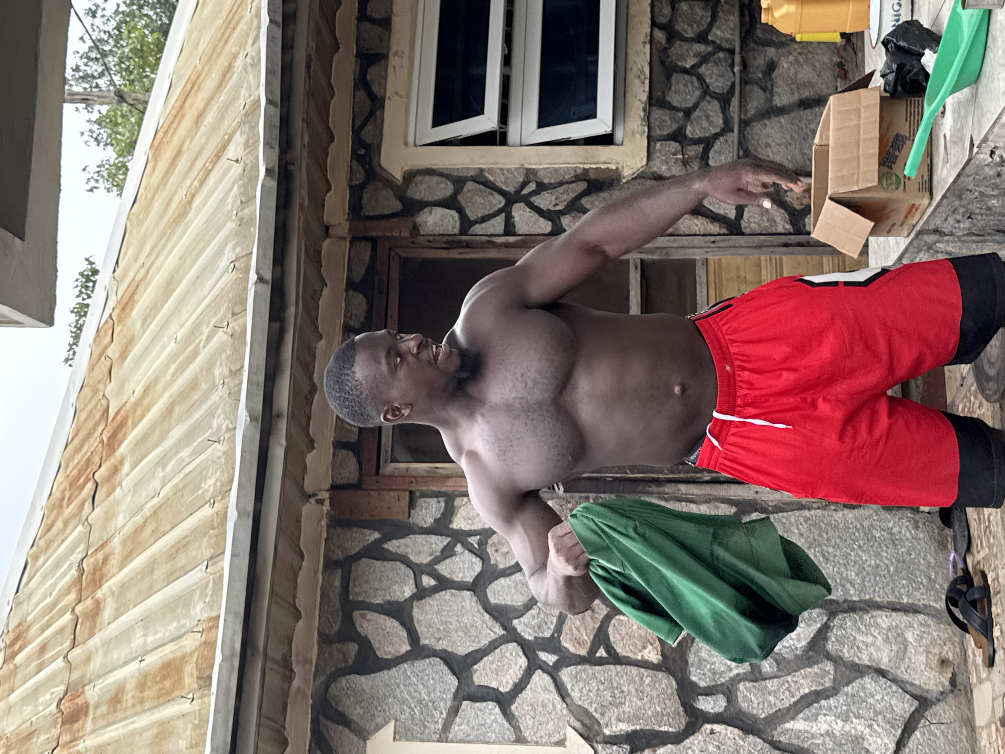 A shirtless Black man wearing red and black shorts stands outside a building with a stone facade and a corrugated metal roof. He is holding a crumpled green garment in his left hand. The man has a muscular physique and is looking to his left. In the background, there is a wooden door, two windows with white frames and tinted glass, and various objects on a concrete surface, including a cardboard box, a green scoop, a black garbage bag, and a yellow plastic container. The ground is paved with stones and there is some greenery visible in the distance. The scene appears to be in a residential or commercial area within the city of Akwanga, Nigeria.