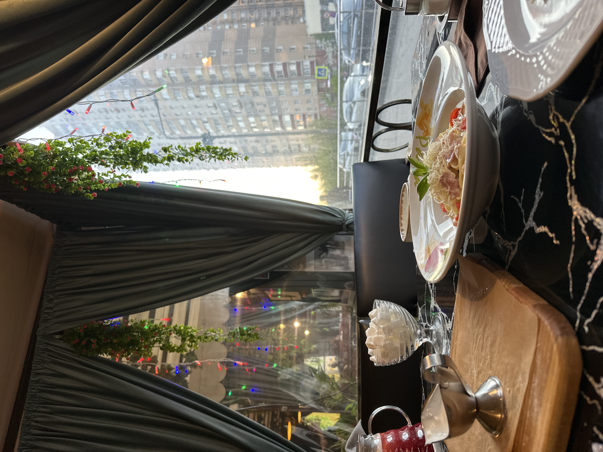 The image shows a restaurant interior with a table set for a meal, viewed through a window displaying a cityscape. The restaurant's interior is decorated with dark green curtains, floral garlands, and multi-colored fairy lights strung across the window frame. On the table, there is a plate of pasta with ham and cheese, along with a smaller dish, a sugar bowl, a cutting board, and a silver dispenser.
Outside the window, a view of apartment buildings under a cloudy or rainy sky is visible. A pedestrian crossing sign is also visible. Given the information, the location is identified as Balashikha, Russia.