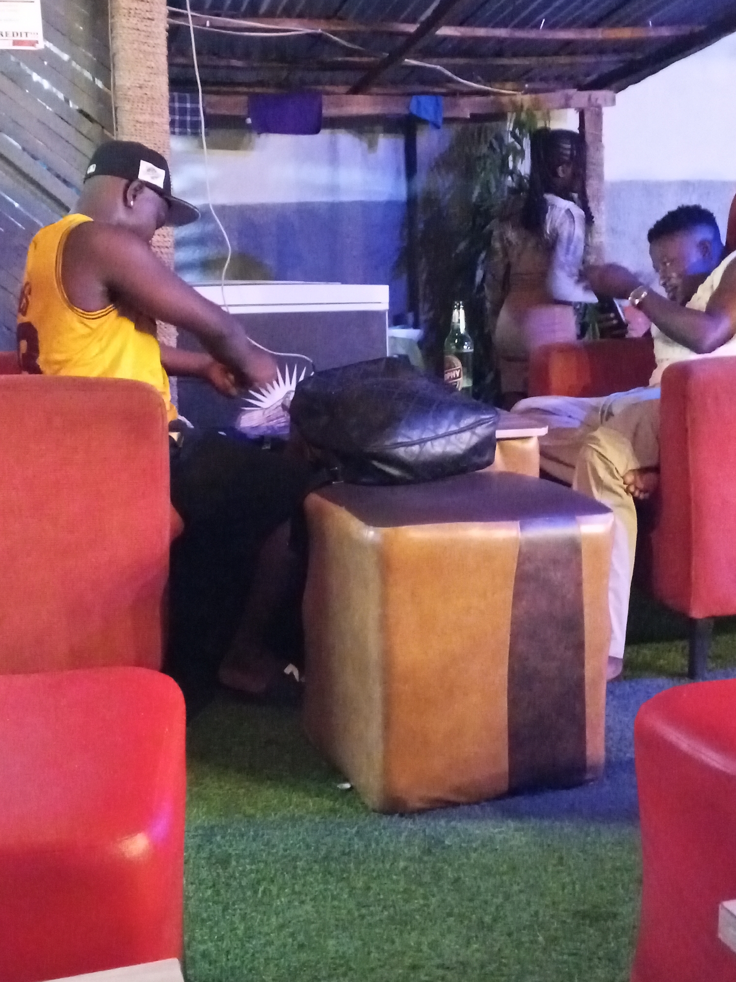 The image shows a dimly lit indoor/outdoor seating area at a bar or restaurant in Adankolo, Nigeria. In the foreground, two men are seated. The man on the left, wearing a yellow basketball jersey and a black baseball cap, is leaning forward with his arms resting on his lap. The man on the right, wearing a light-colored top, is looking down at his phone, which he is holding in his hands. A woman in the background, wearing a light-colored dress, is standing and looking away from the camera. There are several red chairs and a brown ottoman in the seating area. A bottle of what appears to be beer is on a table behind the man on the right. The ground is covered with artificial turf. The lighting is low, and the overall atmosphere appears relaxed.