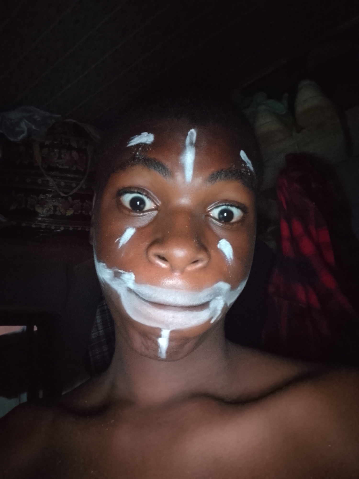 A close-up, dimly lit portrait captures a young individual with dark skin looking directly into the camera. The person's eyes are wide and alert, and a subtle smile plays on their lips. Their face is adorned with distinct white paint patterns. A prominent vertical white line runs down the center of the forehead, flanked by shorter vertical lines above each eyebrow. White lines extend horizontally from the outer corners of both eyes. The mouth is outlined with a semi-circular white stripe, creating an exaggerated smile, and a vertical line extends downwards from the chin. The person's shoulders and upper chest are visible, suggesting they are shirtless or wearing attire that exposes their upper body.
The background is very dark and indistinct, indicating an indoor setting with low ambient light, possibly at night. Faint details suggest the presence of stacked items or textiles, with a patterned material visible on the left and a folded red fabric on the right. The overall impression is one of playfulness or curiosity, with the face paint possibly signifying a cultural ritual, performance, or artistic expression. There is no visible text in the image.