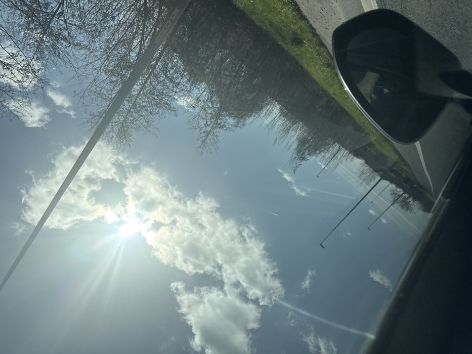 The image captures an outdoor scene on a bright, sunny day, likely viewed from the interior of a vehicle moving along a road in Lokeren, Belgium. The sky, predominantly blue, features scattered white cumulus clouds. The sun is prominently visible in the upper left quadrant, partially obscured by clouds, emitting distinct sunbeams. Multiple linear contrails from aircraft are present across the sky.
To the right, a dense line of bare deciduous trees and brush borders a patch of green grass, indicative of late winter or early spring. A dark, tall pole, likely a streetlight or utility pole, extends diagonally from the middle-right to the upper center, traversing the sky. Below, a paved road with a solid white edge line is visible. Several dark, slender streetlights are evenly spaced along the roadside, extending into the distant perspective.
In the lower right foreground, a portion of a dark car's interior, including the top edge of a dashboard or door panel, and the black casing of a passenger-side rearview mirror are visible. The mirror's reflective surface shows a blurred, dark reflection. No individuals are depicted.