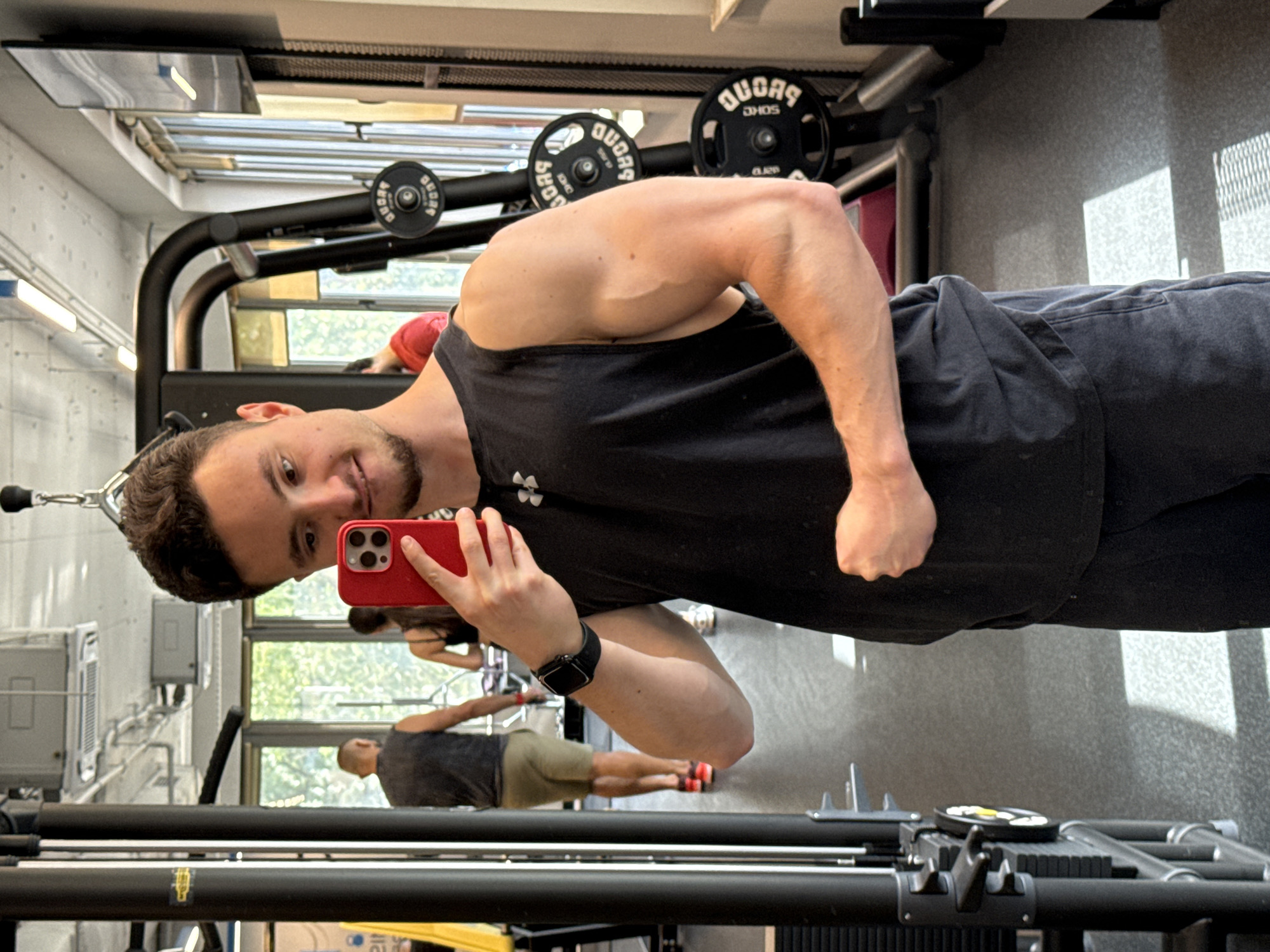 A man is taking a selfie in a gym. He is holding a red smartphone in his right hand and wearing a black tank top and dark gray pants. He also has a black smartwatch on his left wrist. He is in front of a mirror, and the reflection shows other people in the gym. The gym has workout equipment and windows with a view of greenery. The city is Sofia, Bulgaria, but there is no visual evidence in the photo to confirm that location, so it is not possible to confirm the location forensically.
