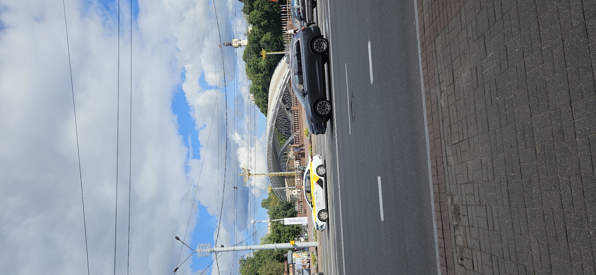 The image shows a street scene in Vitsebsk, Belarus. In the foreground, a white and yellow taxi is driving on the road, followed by a dark gray SUV. In the background, there is a large, modern structure with a curved roof, possibly a stadium or concert hall. Behind that, trees and a clock tower are visible. The sky is partly cloudy with blue patches. The street is paved with asphalt and lined with cobblestones on the sidewalk. Electrical wires are strung across the sky. Several people are walking on the sidewalk in the background.