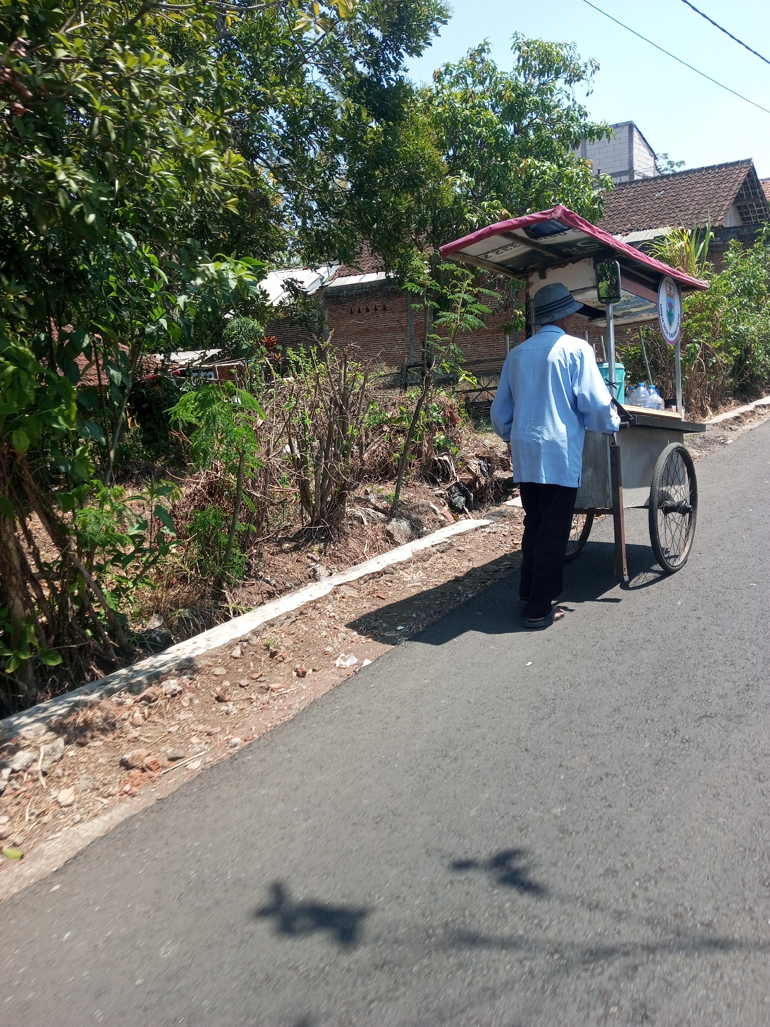 A person wearing a light blue long-sleeved shirt and dark pants, with a grey hat, walks from left to right, pushing a wheeled cart. The cart has a red awning and appears to be a food or drink vendor's cart, with bottles visible inside. The scene takes place on an asphalt road next to a dry, overgrown ditch. In the background, there are brick buildings with tiled roofs and lush green trees. The sky is clear and blue, indicating a sunny day. The location is identified as the city of Purwosari Klampok, Indonesia.