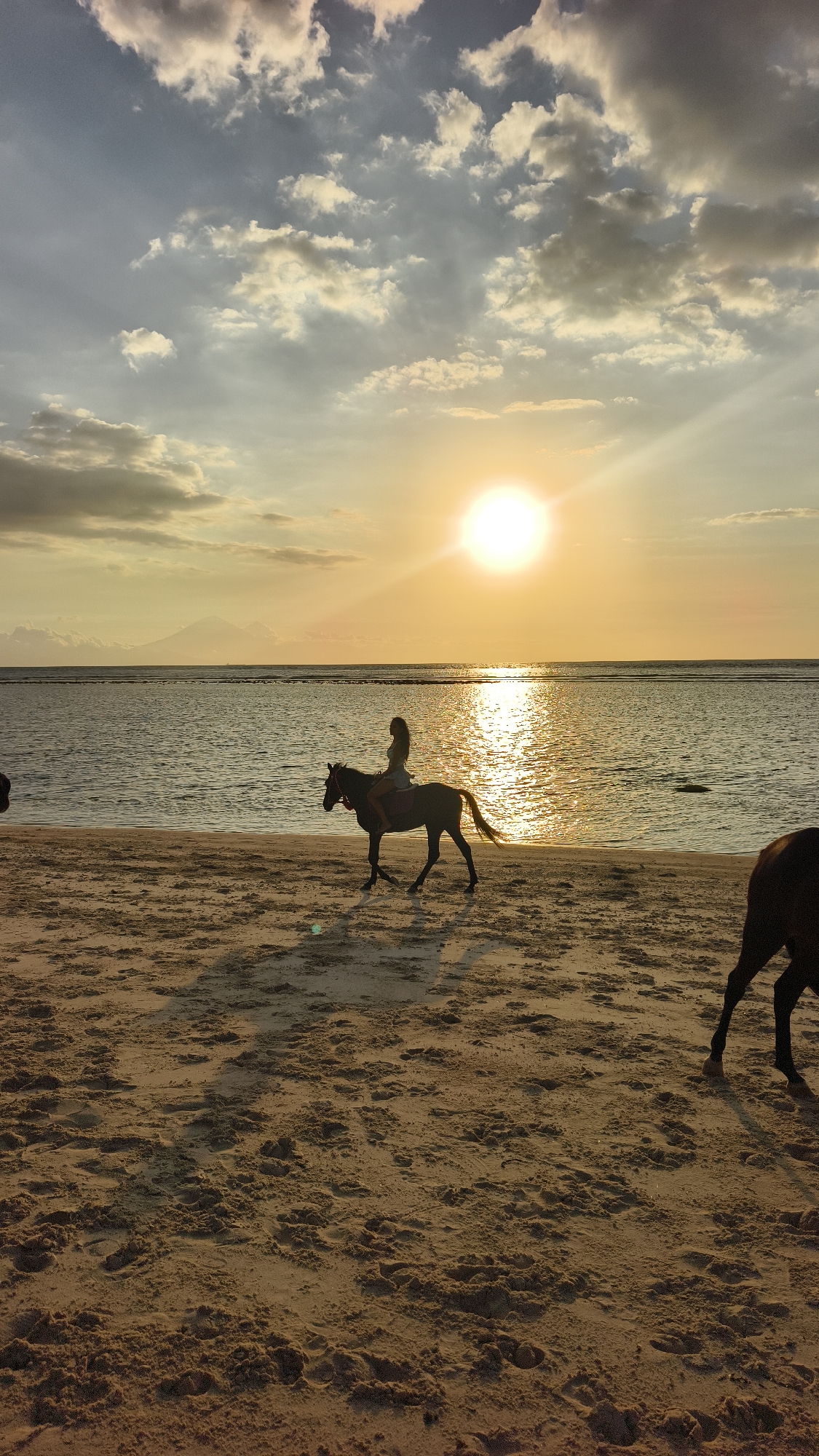 A photograph captures a beach scene at sunset. A person sits on a dark horse that is moving from left to right in the frame. The sun sits high in the sky, reflecting light on the water. To the right of the horse is the rear of another horse that is moving out of the frame. The sand is brown and textured, and the horizon is visible in the middle of the image. The location is Unknown, Indonesia.