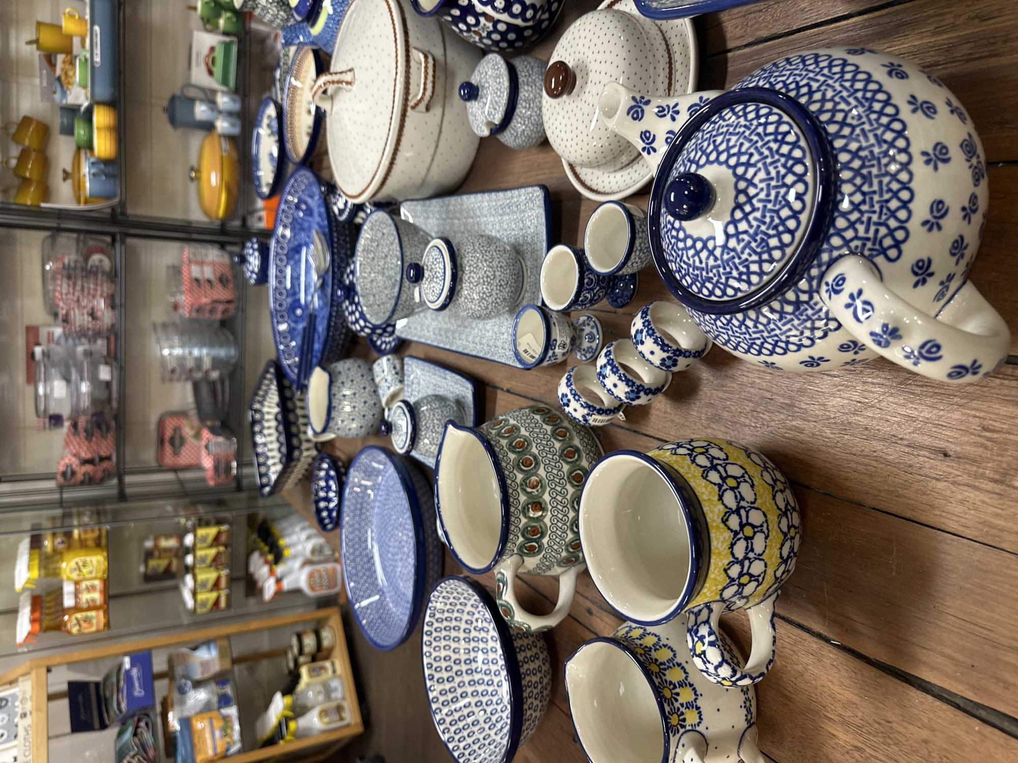 This image depicts a display of Polish pottery, likely in a shop, with a focus on ceramic tableware. The pottery features intricate blue and white floral patterns, as well as some pieces with yellow floral accents. Visible items include teapots, mugs, bowls, serving dishes, a butter dish, and egg cups. The pottery is arranged on a wooden table, with shelves of other merchandise visible in the background, suggesting a retail setting. There are no people present in the image. The scene is indoors, with wooden flooring and shelving units. The lighting suggests ambient indoor lighting. The overall impression is one of a curated collection of decorative and functional ceramic items. The specific location in Bellingen, Australia, is not deducible from the image itself.