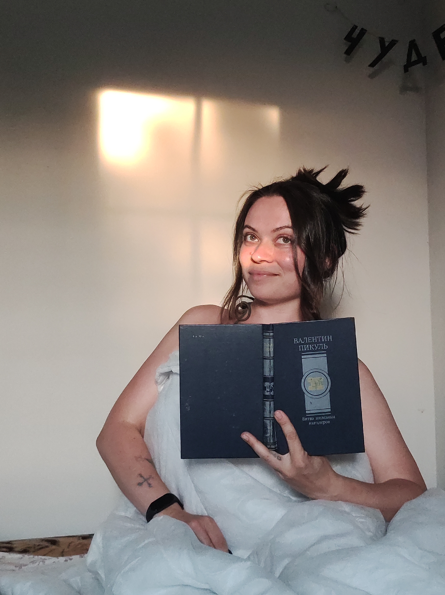 A woman is sitting in bed, holding an open book in front of her. She is partially covered by a white comforter or blanket. She has dark hair, styled in a messy bun, and is looking slightly upwards. Her right arm has a cross tattoo. The book's cover is dark with an image of a watch and text in Cyrillic script. There is a square of sunlight on the wall behind her. Above her head are several black Cyrillic letters. The location is an interior setting.