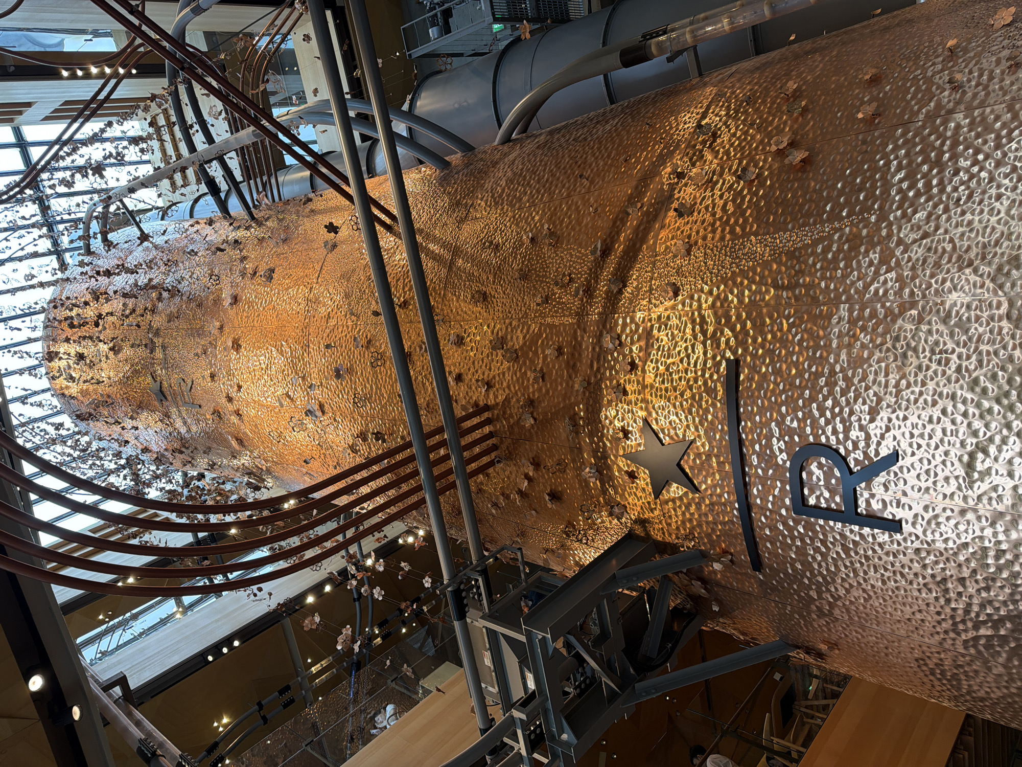 The image shows the interior of the Starbucks Reserve Roastery in Meguro, Japan. The focal point is a large, copper-colored roasting tower that dominates the center of the frame. The tower is textured with a hammered finish and adorned with numerous small, metallic flower-like ornaments that appear to be falling or attached to the surface. Above the tower, a complex network of pipes, metal structures, and decorative elements made of copper tubing and delicate chains with flower ornaments are visible. Natural light streams in from a large skylight at the top, illuminating the scene. A large black Starbucks logo, consisting of a star and an 'R', is prominently displayed on the lower section of the roasting tower. In the background, balconies with wooden railings and seating areas are visible, along with glimpses of the surrounding interior architecture, including wooden beams and more lighting fixtures. Two individuals are partially visible on a balcony in the upper left portion of the image. The overall impression is of a large, industrial yet artistically designed space, indicative of a flagship store or a unique architectural experience.
