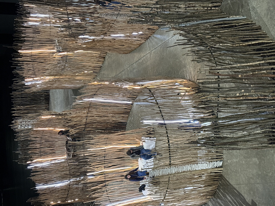 This image depicts an immersive art installation within The Curve gallery at the Barbican Centre, London, United Kingdom. The scene features a labyrinthine structure constructed from numerous upright, slender wooden sticks or branches, forming curving pathways and circular enclosures on a smooth, light grey concrete floor. Interspersed vertically among the wooden elements are luminous white light tubes, some emitting a cooler white light and others a warmer, amber glow, creating alternating bands of light and shadow along the walls of the maze. The ceiling and background are dark and indistinct.
In the foreground-left, two individuals are seated on a white, fabric-covered bench or platform within one of the circular enclosures. One person wears a dark blue top and blue jeans, while the other wears a grey jacket and blue jeans. A dark bag rests on the platform beside them. A segment of the enclosure's wall directly in front of them is composed of thin, light-colored horizontal elements. Further into the installation, in a separate, elevated circular area, a third individual with dark hair, wearing dark clothing, is partially visible, also seated on a white platform. A fourth person's lower body is faintly discernible behind a stick wall on the right side of the frame. All individuals appear to be observing or resting within the installation.