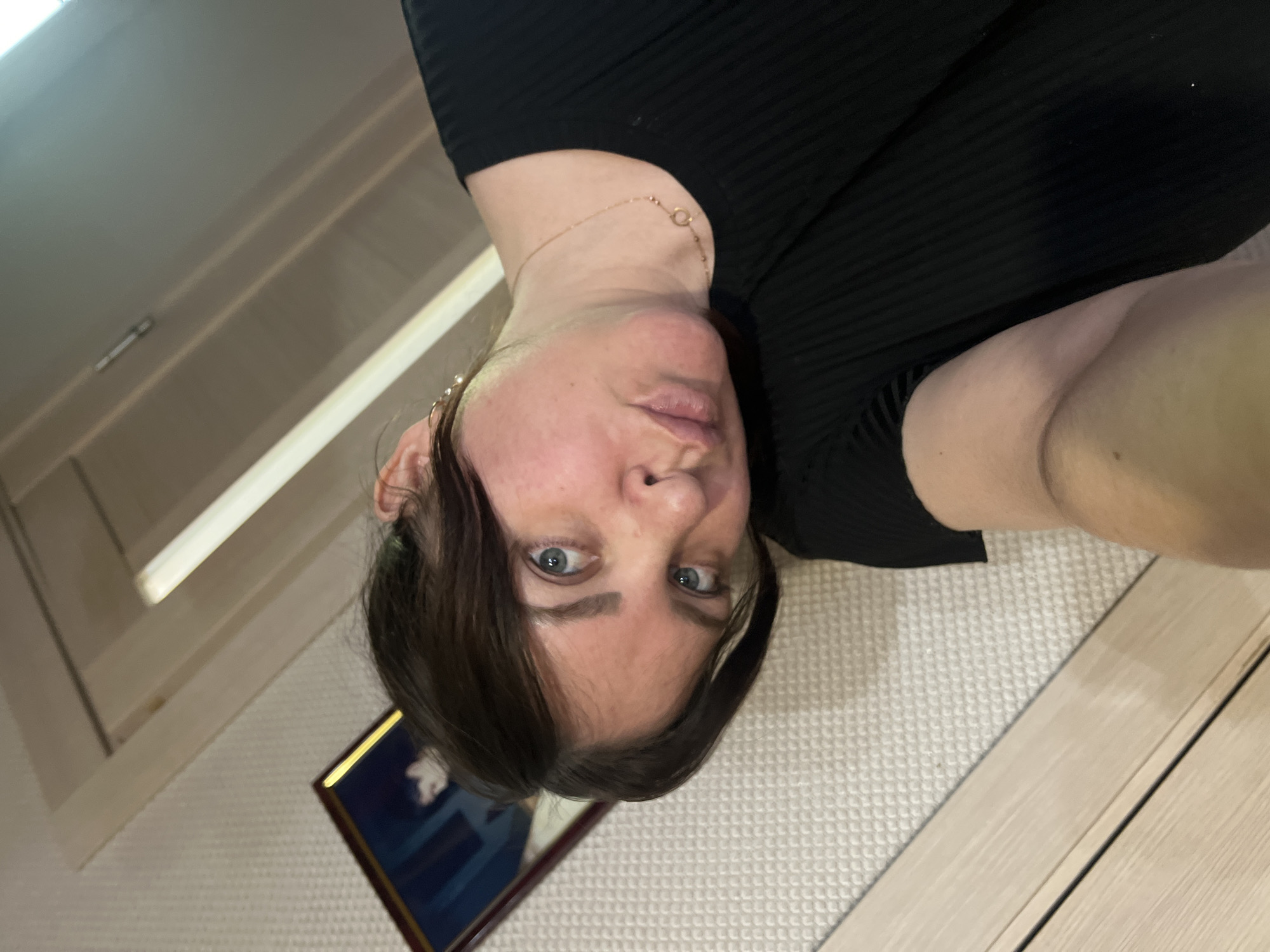 A photograph shows a woman indoors, with her face in the lower center of the frame. She has fair skin, dark hair with light highlights, and blue eyes. She wears a black ribbed garment. A gold necklace is visible around her neck.
The background includes a textured wall, a section of a door, a framed picture hanging on the wall, and a light fixture on the ceiling. The location is unspecified, and there is no clear indication in the image that places it in Noviy, Russia.