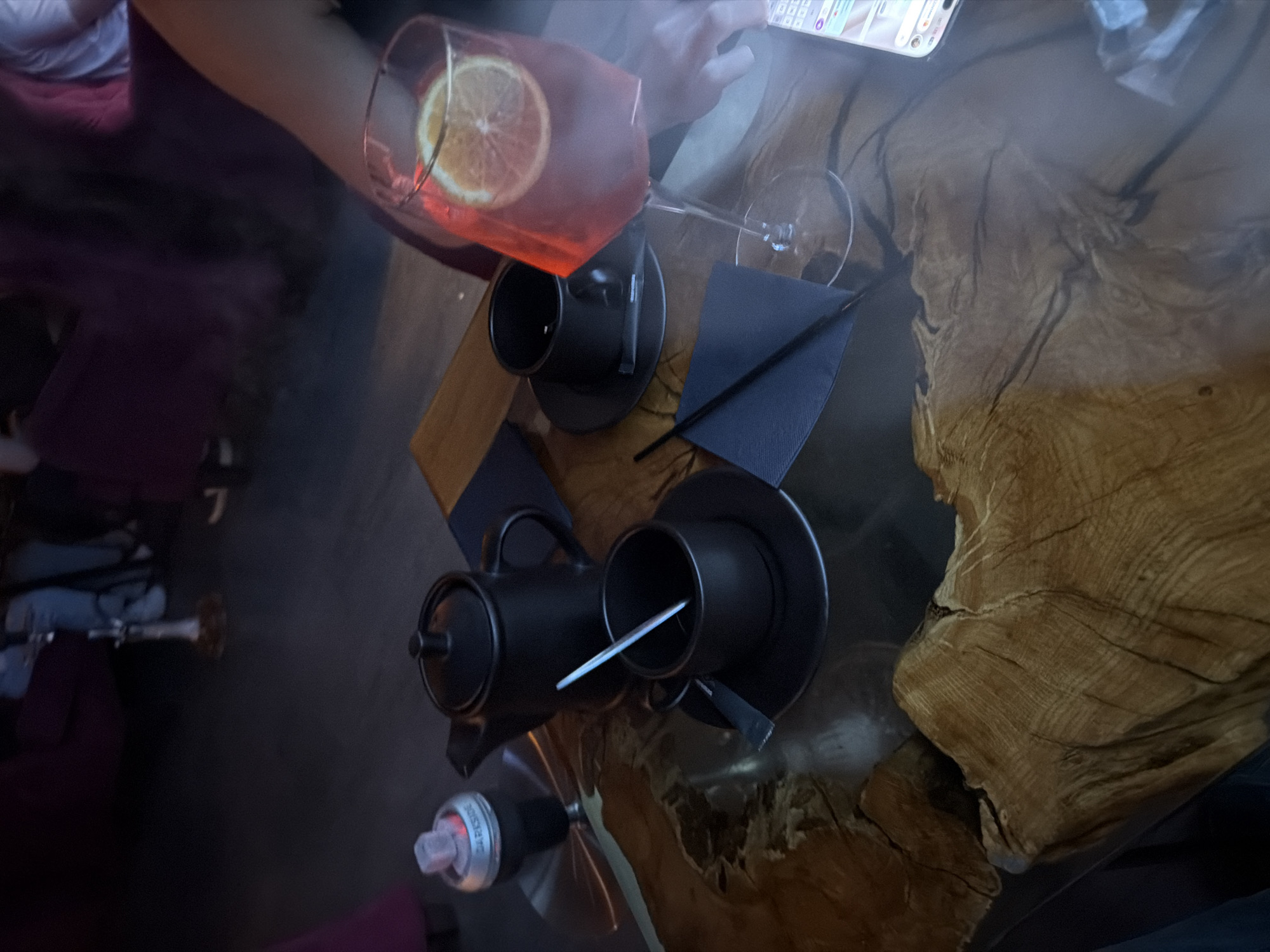 The image shows a table setting, including tea service and a cocktail, likely in Krasnoyarsk, Russia. The table is a rough-hewn, varnished tree trunk section. A teapot and teacups are present, along with a cocktail glass containing a red drink and a slice of citrus. A wine glass and napkins are visible. An arm holding a cell phone is partially visible above the table. A hookah is present next to the teapot. The lighting is low.