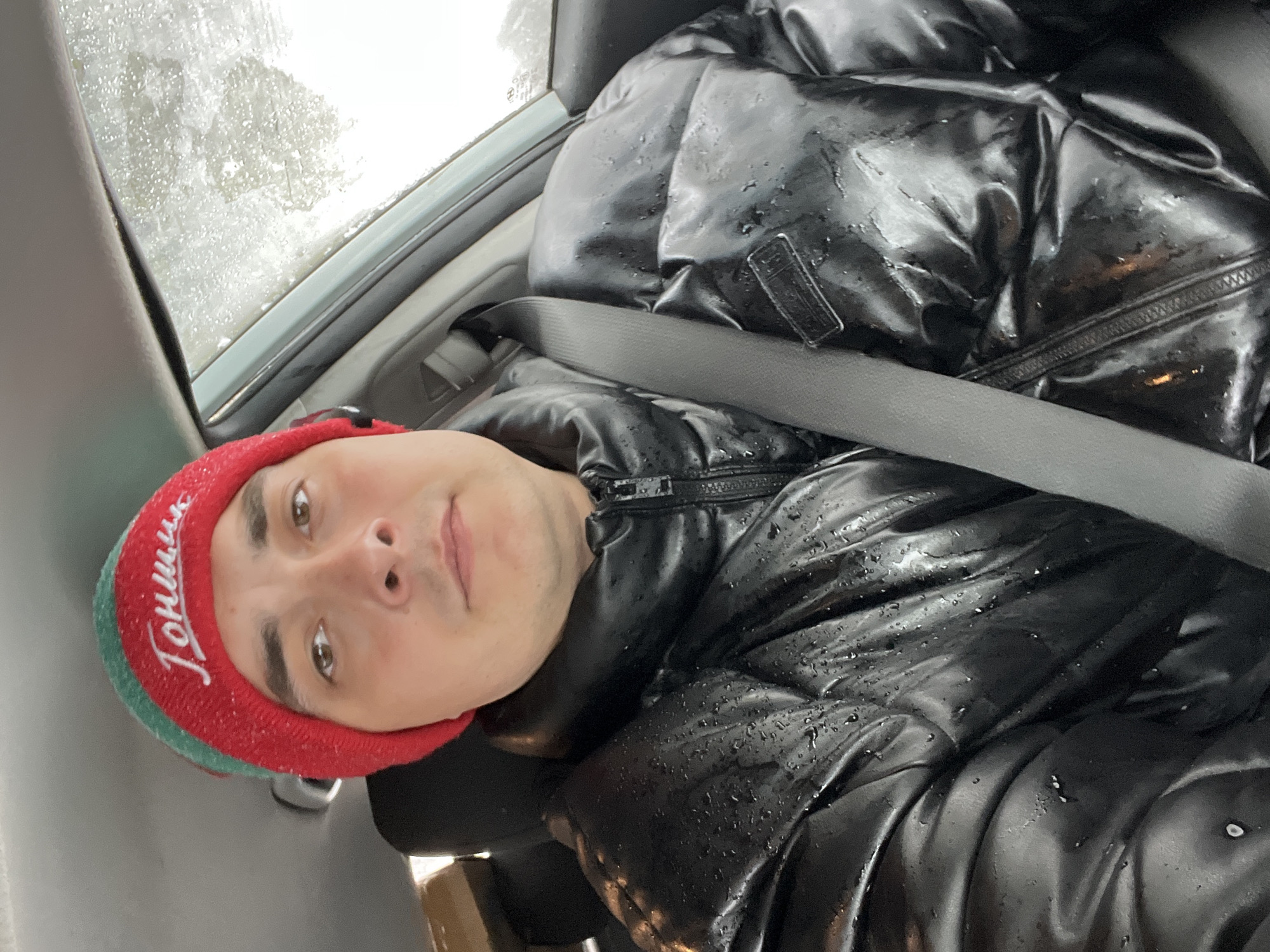 A man is sitting in the passenger seat of a car, wearing a shiny black puffer jacket and a red and green hat with the word "Гонщик" (Racer) written on it. He is looking at the camera. The seatbelt is fastened across his chest. Rain or snow is visible on the car window in the upper left corner of the image. The location is likely Razdolye, Russia based on the hat inscription.