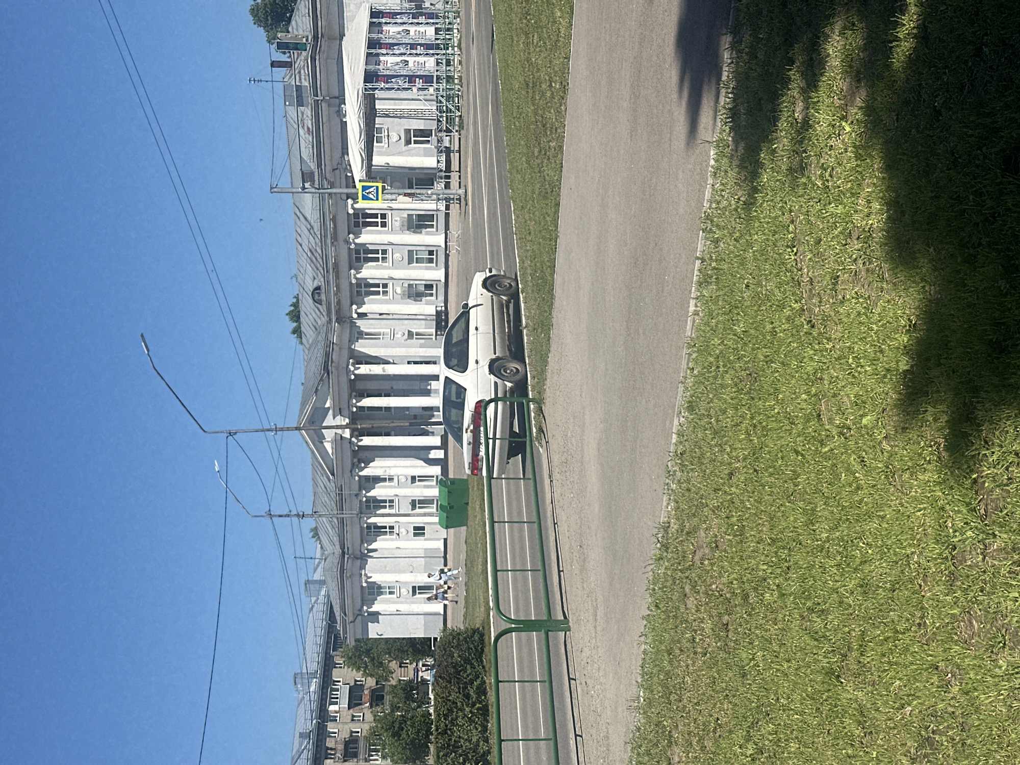 This image depicts a bright, sunny day in Novoaltaysk, Russia. In the foreground, a patch of vibrant green grass borders a paved area. A low, green metal fence runs parallel to a road, with a white car parked partially on the asphalt to the right of the fence. The car appears to be a sedan, showing signs of wear. Beyond the road, a large, white, neoclassical building with a colonnade and a flat roof dominates the background. To the left of this main structure, a taller, gray apartment building with multiple windows is visible. Several people are walking on a sidewalk in front of the neoclassical building, and two large green trash bins are placed near the building's entrance. A pedestrian crossing sign with a yellow triangle and a figure walking is also present. Overhead, power lines stretch across a clear blue sky, and a lamppost stands near the right side of the frame. The scene suggests a public space within the city, likely near a cultural or administrative center.