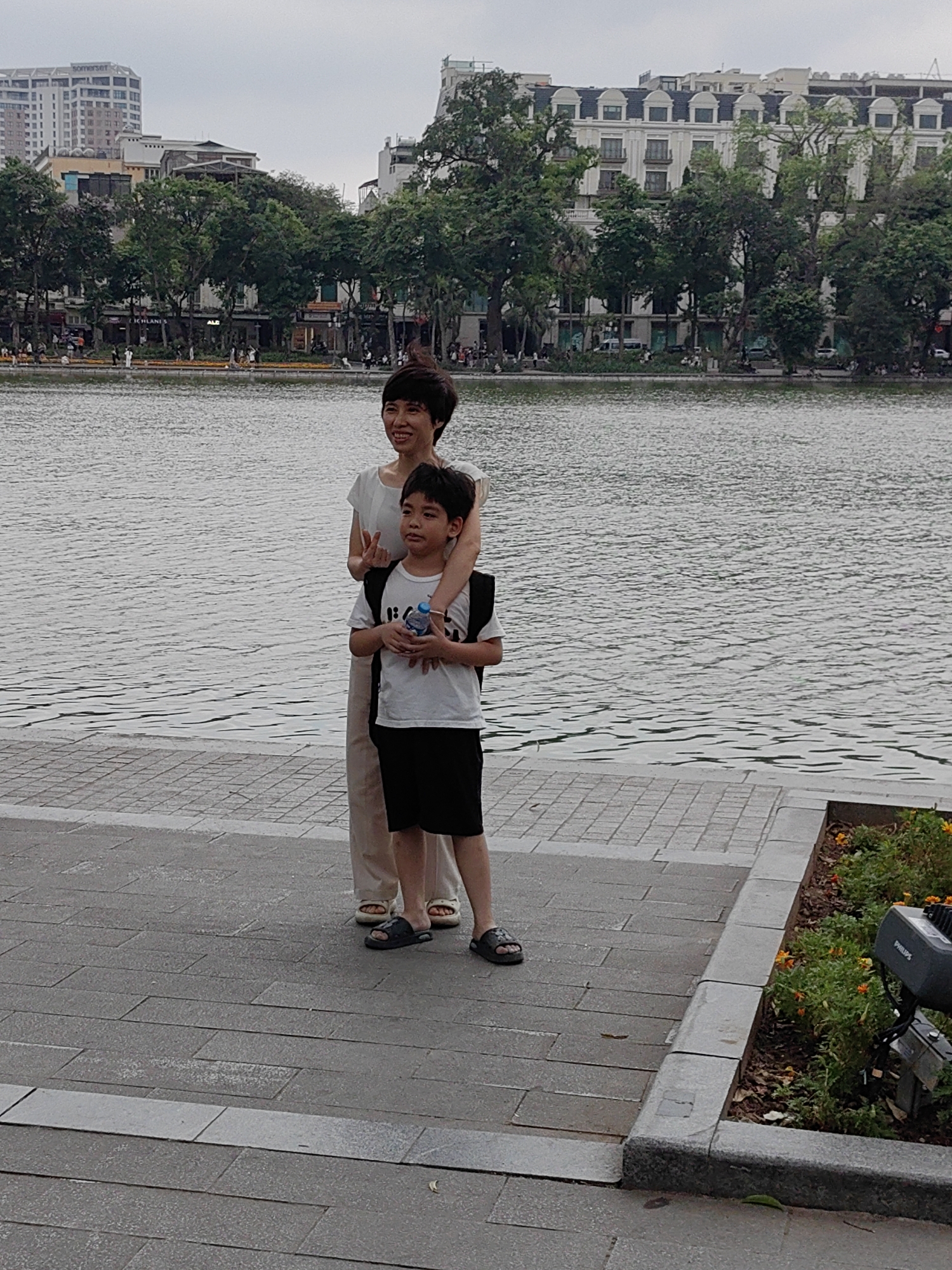 An outdoor, eye-level shot captures a woman and a boy standing beside a body of water, likely a lake, on a paved promenade in Hà Nội, Vietnam. The woman, positioned slightly behind the boy, has her right arm draped over his shoulder. She has short, dark hair and is wearing a light-colored top and light-colored pants, with open-toed sandals. Her face shows a gentle smile, and she is looking towards the left, off-camera.
The boy, who appears to be pre-teen, stands in front of the woman, looking ahead with a slightly serious or thoughtful expression. He is wearing a white t-shirt with a graphic design, black shorts, and dark slide sandals. He holds a clear plastic water bottle in his hands and wears a black backpack.
Behind them, the calm water extends across the middle ground, reflecting the overcast sky. On the far bank of the lake, various buildings and lush green trees are visible. One prominent building in the distance on the left features the text "SOMERSET" on its roof. Other buildings of varying architectural styles line the waterfront, interspersed with dense foliage. A paved walkway, similar to the foreground, is visible on the opposite side, with small figures of people suggesting activity.
The immediate foreground features a grey, paved stone walkway, partially obscured by a decorative planter box filled with green plants and small orange flowers. A black outdoor light fixture with the brand "PHILIPS" visible on its side is positioned within this planter box on the right. The overall impression is a daytime scene under an overcast sky, with the subjects posing by the water in an urban setting.