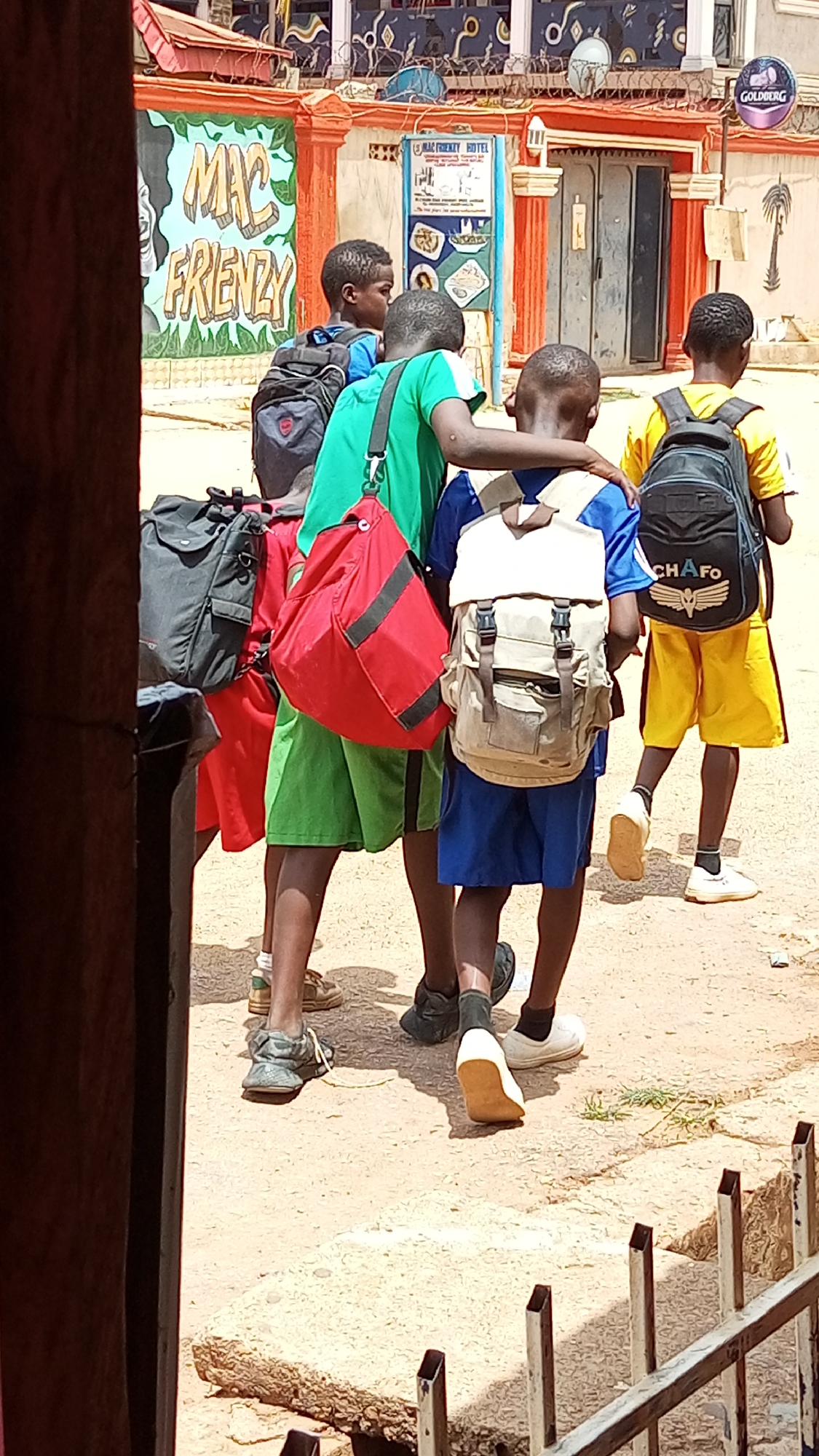 Four school-aged boys are walking along a dirt path in Sabon Tasha, Nigeria. Each boy is wearing a different color uniform and backpack. The boy in green has his arm around the boy in blue. Behind the boys is a building with colorful graffiti and a sign that reads "Mac Friendly Hotel." The environment appears to be an outdoor setting.