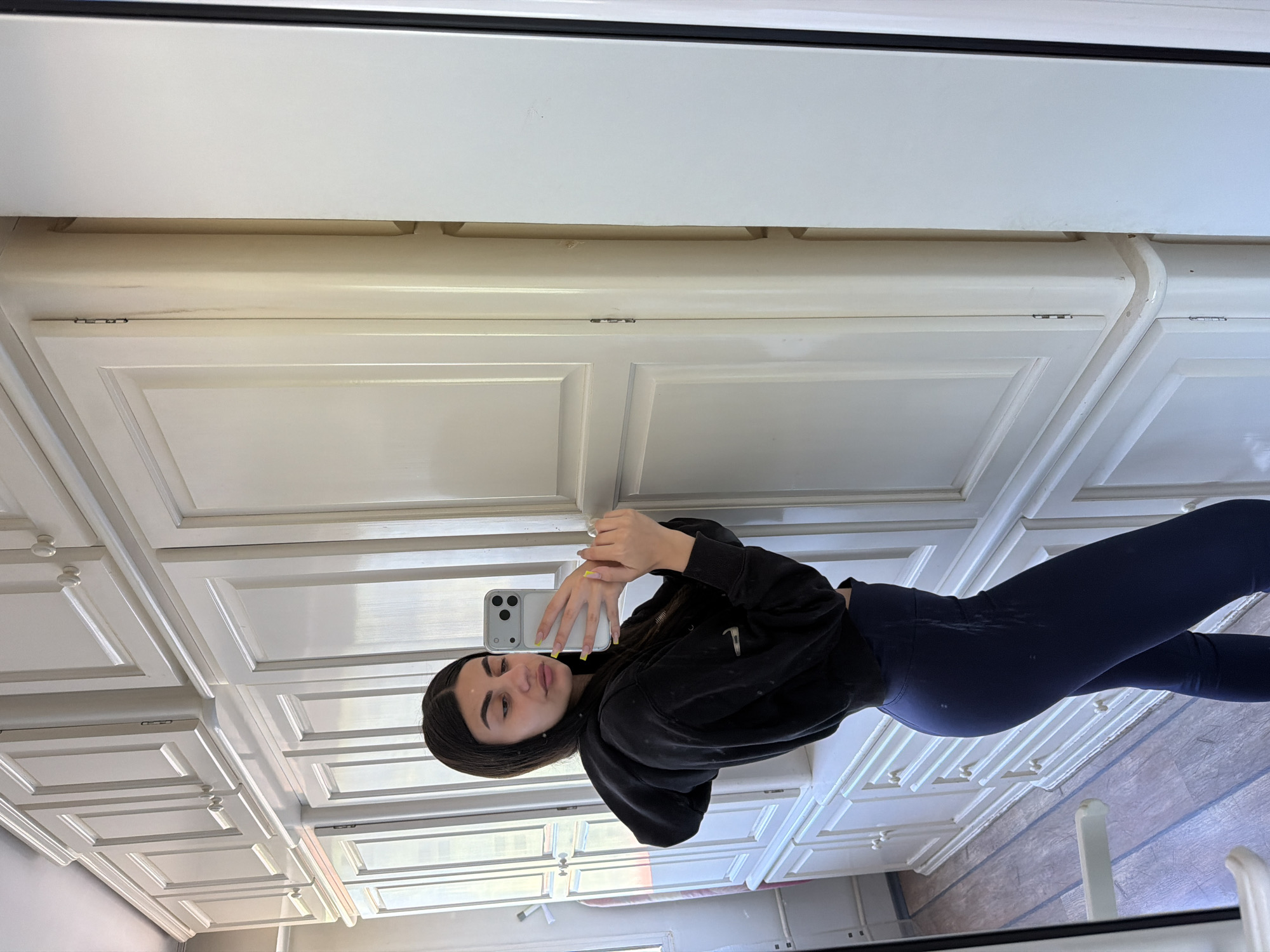 A woman is taking a mirror selfie in a bathroom with white cabinets. She is wearing a black sweatshirt and blue leggings. Her hair is dark. She is holding a white smartphone in her hands. The bathroom floor appears to be wooden. The location cannot be pinpointed to Gabrovo, Bulgaria from the provided information.