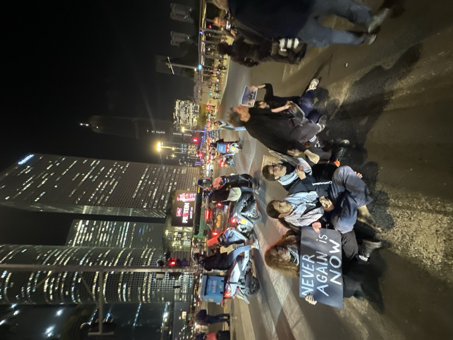 A group of people is lying on the road, possibly blocking traffic at night, holding a sign that reads "Never Again Is Now". Skyscrapers lit with many windows rise in the background. There are scooters and stationary cars around them. Some individuals are wearing keffiyehs.