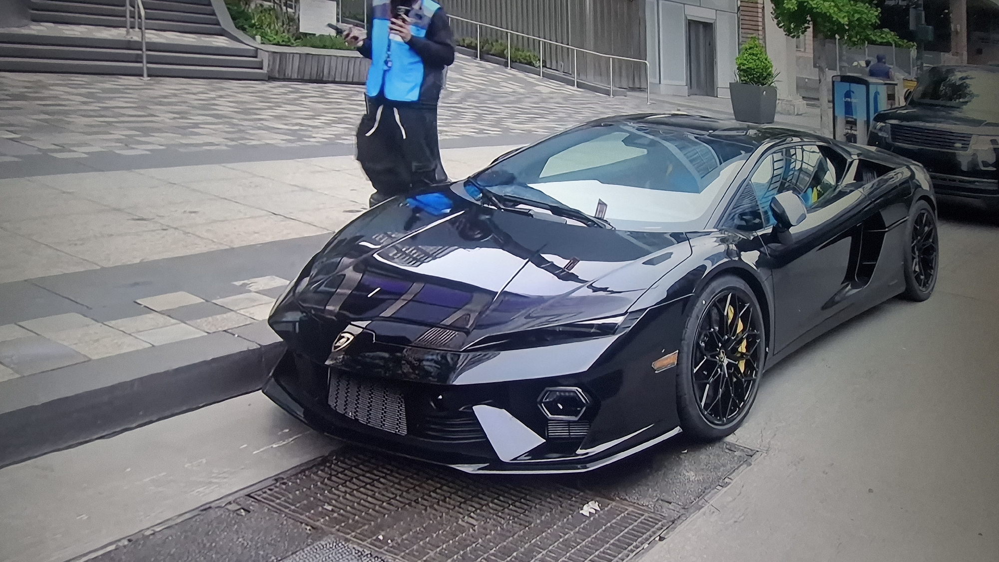 A black Lamborghini sports car is parked on a city street next to a grated storm drain. A person wearing a blue vest and dark clothing stands near the front of the car, holding a device, possibly a phone or electronic tablet. Behind the car, another vehicle, appearing to be a black SUV, is partially visible. There are buildings in the background, including one with steps leading up to it. The setting suggests an urban environment of unknown location.