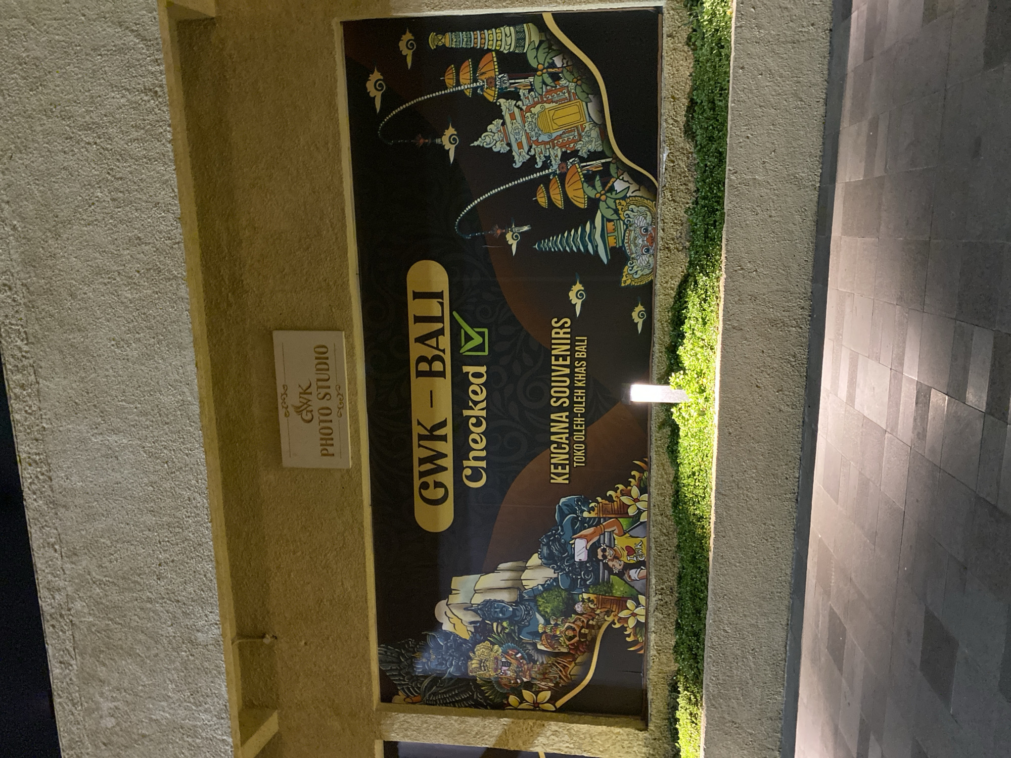 This media file depicts a sign for "GWK PHOTO STUDIO" and "KENCANA SOUVENIRS TOKO OLEH-OLEH KHAS BALI" in Kuta Selatan, Indonesia. The sign is large and features intricate Balinese artwork, including depictions of temples, mythical creatures, and people. The artwork is vibrant and detailed, with a dark background that makes the colorful elements stand out.
To the left of the main sign, there is a smaller, rectangular sign that reads "GWK PHOTO STUDIO". This sign is white with black text and a small graphic. The main sign has large, bold text that says "GWK - BALI" and "Checked ✓", suggesting a checkmark of approval or verification. Below this, the text "KENCANA SOUVENIRS" and "TOKO OLEH-OLEH KHAS BALI" indicates that it is a souvenir shop selling typical Balinese items.
The scene appears to be outdoors, with a paved walkway and lush green shrubbery visible to the right of the sign. The lighting suggests it could be evening or night, as a light fixture is visible above the greenery. The overall impression is one of a tourist-oriented establishment in Bali, showcasing local culture and offering souvenirs. The artwork implies a rich cultural heritage and potentially a lively atmosphere for visitors.