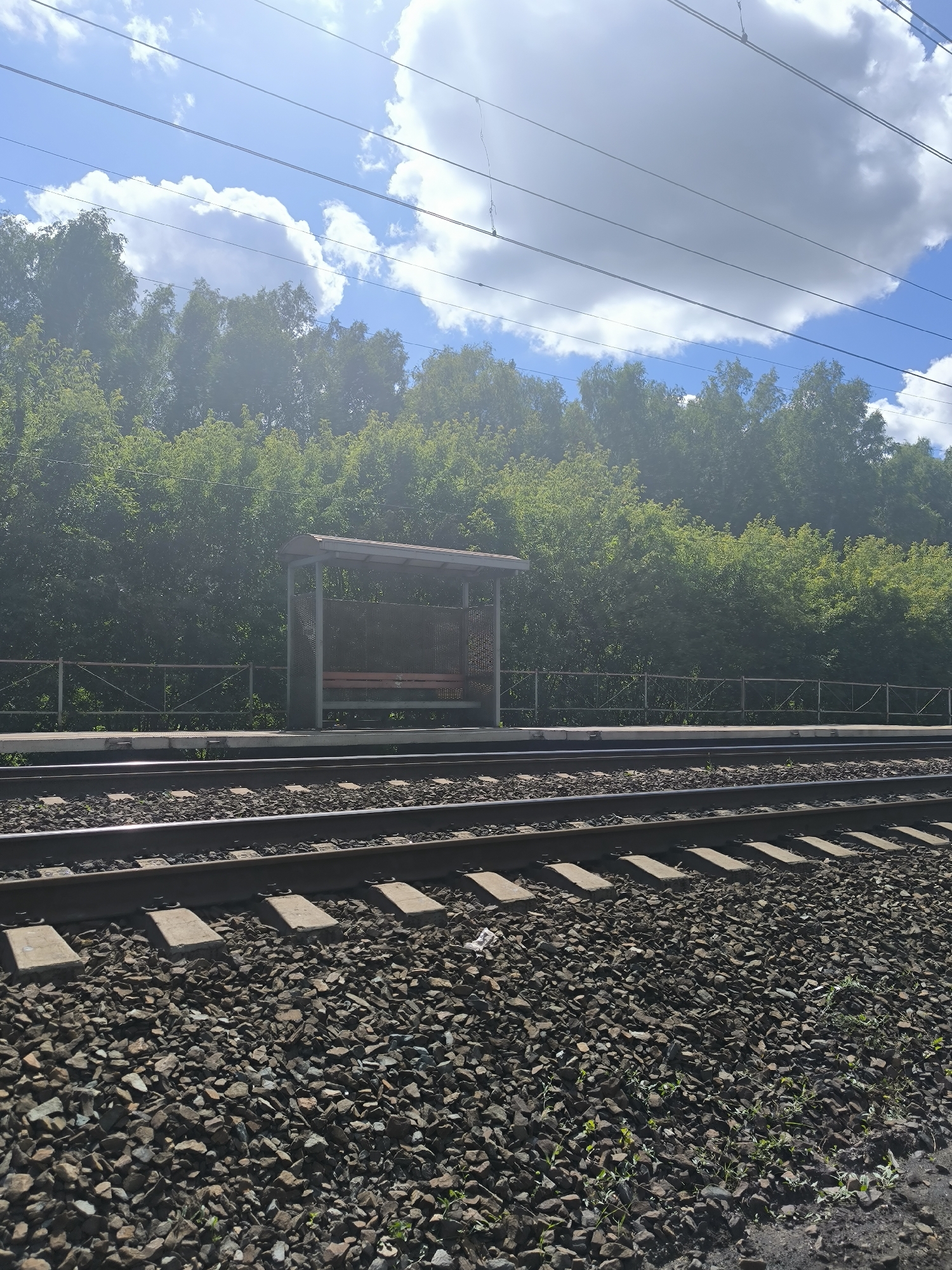 The image shows a train station platform with railway tracks in the foreground. A bus shelter with a bench is visible on the platform, behind a metal fence. A dense line of green trees forms the background, with a bright blue sky and white clouds above. Overhead power lines are also visible. The scene is devoid of people. The location is identified as Maiskii, Russia.