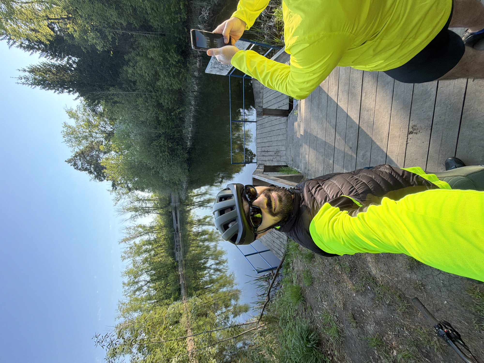 A man wearing a cycling helmet and sunglasses poses for a photo on a dock by a river or lake. He is dressed in a neon green jacket and a black vest. Another person, wearing a similar neon green jacket and black shorts, holds a smartphone to take the photo. The dock is made of wooden planks and has a blue railing at the end. Trees and greenery surround the body of water. The sky is blue. The scene appears to be in a park or recreational area. While the exact location in Tyumen cannot be determined from this image alone, the presence of the dock, the trees, and the river indicate that it could be on the Tura river.