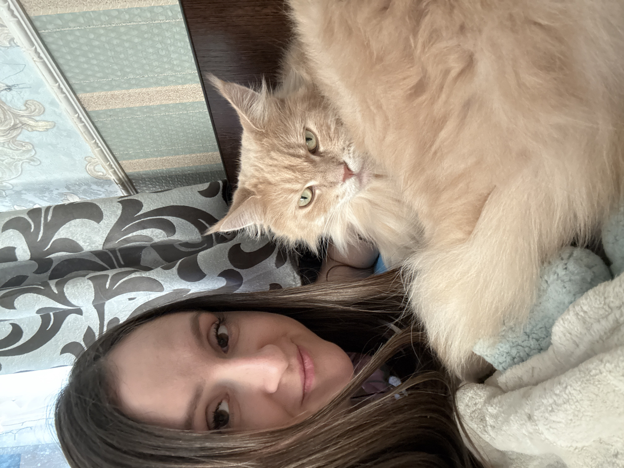This is a close-up, high-angle, selfie-style photograph of a woman and a cat in Kroyanskoye, Russia. The woman, with long brown hair, is positioned on the left side of the frame, looking directly at the camera with a slight smile. She has brown eyes and is wearing makeup. Beside her, on the right, a fluffy, light orange cat with green eyes is looking off to the side with a somewhat stern expression. The cat's fur is dense and long. They are both lying down on soft, light-colored bedding, possibly a blanket and a bolster. In the background, a curtain with a dark floral pattern is visible, as well as striped wallpaper and a wooden surface. The overall impression is one of domestic comfort.