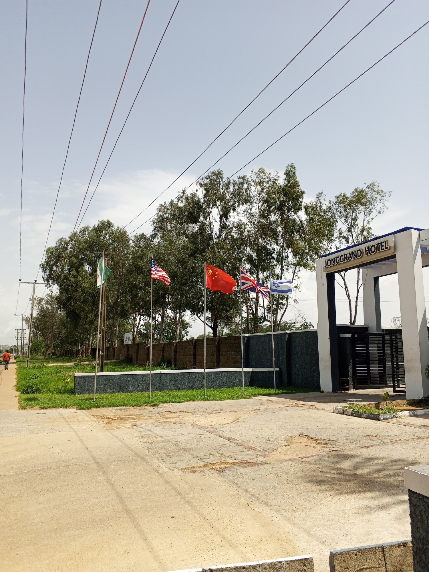 The image depicts the entrance to the Jonggrand Hotel in Bukuru, Nigeria. Several national flags are displayed in front of the hotel: Nigeria, the United States, China, the United Kingdom, and Israel. The hotel entrance features a white structure with the hotel name prominently displayed in dark lettering. A black metal gate and security fencing are visible. The surrounding environment includes a paved area in the foreground, grass, and trees. In the background, a person walks along a sidewalk on the left side of the image. Overhead, power lines are strung across the sky.