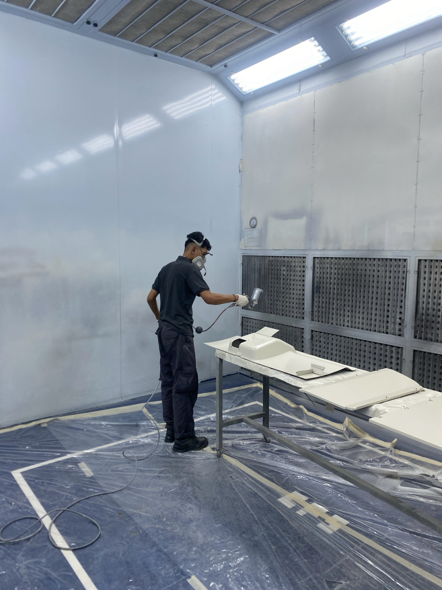 A person, dressed in dark work clothes, a respirator mask, and gloves, is spray painting a light-colored object in a spray booth. The booth has white walls and ceiling with fluorescent lighting. A metal table holds the object being painted, and the floor is covered with blue plastic sheeting and white tape. The background shows industrial ventilation filters. The location is a spray booth in Batam, Indonesia.