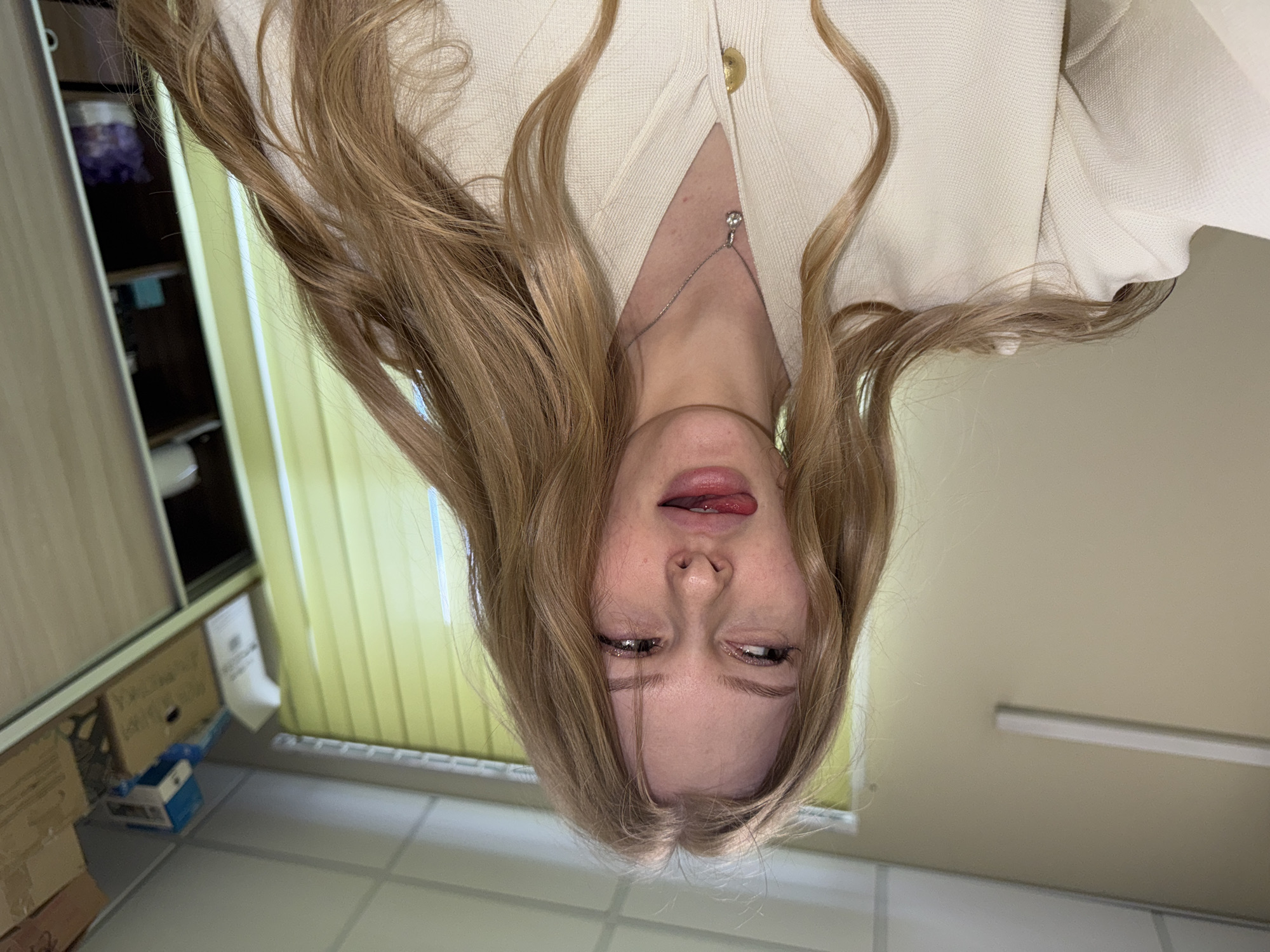 The image is an inverted selfie of a young woman with long, blonde hair. She is wearing a light-colored cardigan and a delicate necklace. Her expression is playful, with her tongue sticking out and eyes squinting.
The background suggests an indoor setting, possibly an office or storage area. There is a cabinet or shelving unit visible on the left, with boxes stacked on the floor. A window with closed, vertical blinds is in the background. The ceiling has a fluorescent light fixture.
There is no definitive information in the image to indicate the location is in Perm, Russia.