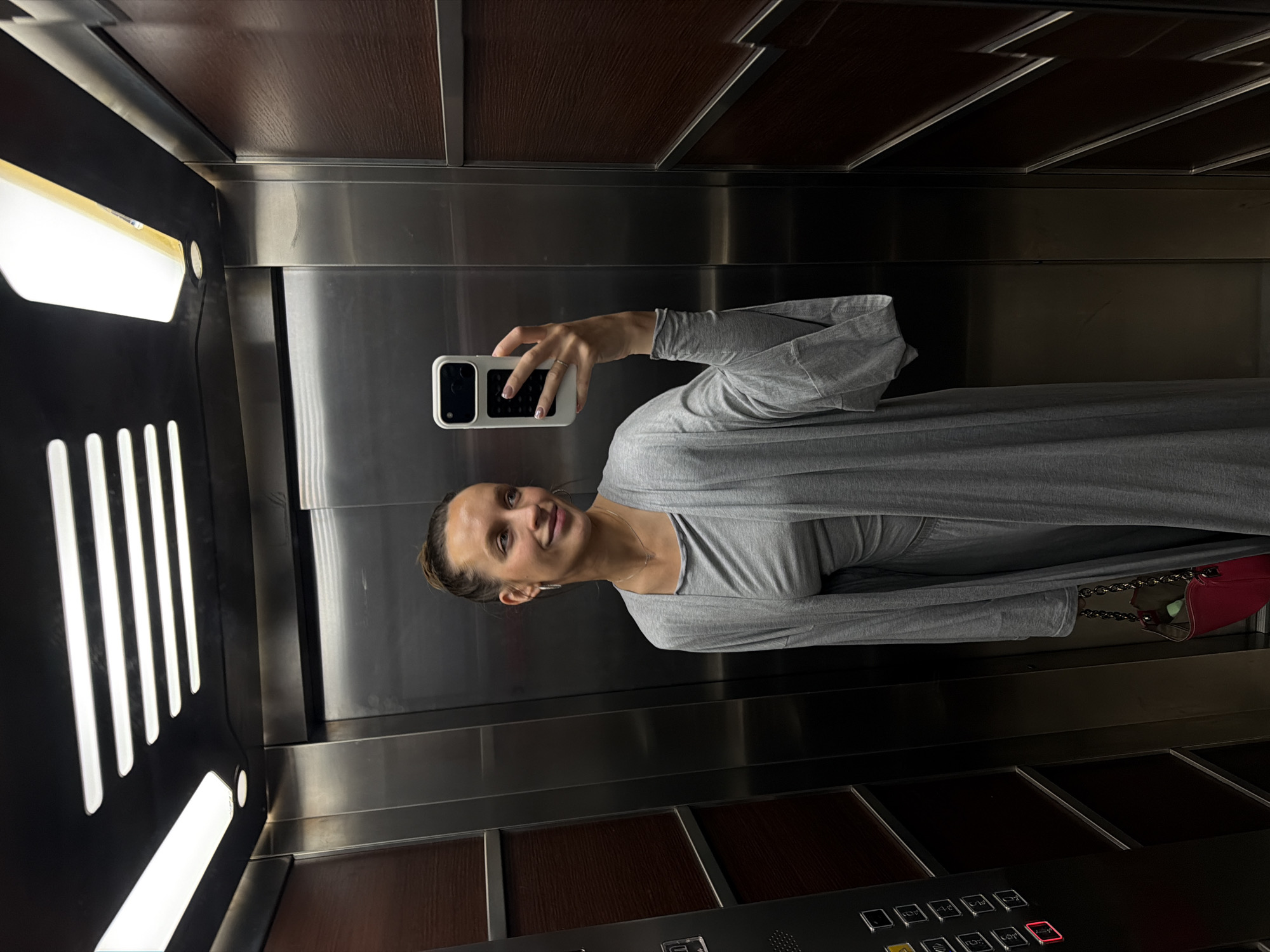 An image captures a woman taking a selfie inside a modern elevator in Budva, Montenegro. The woman is positioned on the right side of the frame, smiling broadly and looking at her smartphone, which she holds in her right hand. She is wearing a long-sleeved, flowing gray dress or top. A small, red handbag with a metallic chain strap hangs from her left side.
The elevator interior is characterized by reflective metallic panels, likely brushed stainless steel, alternating with dark wooden panels on the ceiling and possibly the lower sections of the walls. Bright vertical light panels are visible on the left side of the elevator, casting reflections on the polished surfaces. At the bottom of the frame, an elevator control panel is visible, featuring multiple square buttons. Text visible on these buttons includes "TOP," "LOFT," "ALARM," and "CALL." One of the floor selection buttons, possibly "LOFT," is illuminated in red, indicating a selection. The overall lighting within the elevator is bright and even. The woman appears cheerful and relaxed.
